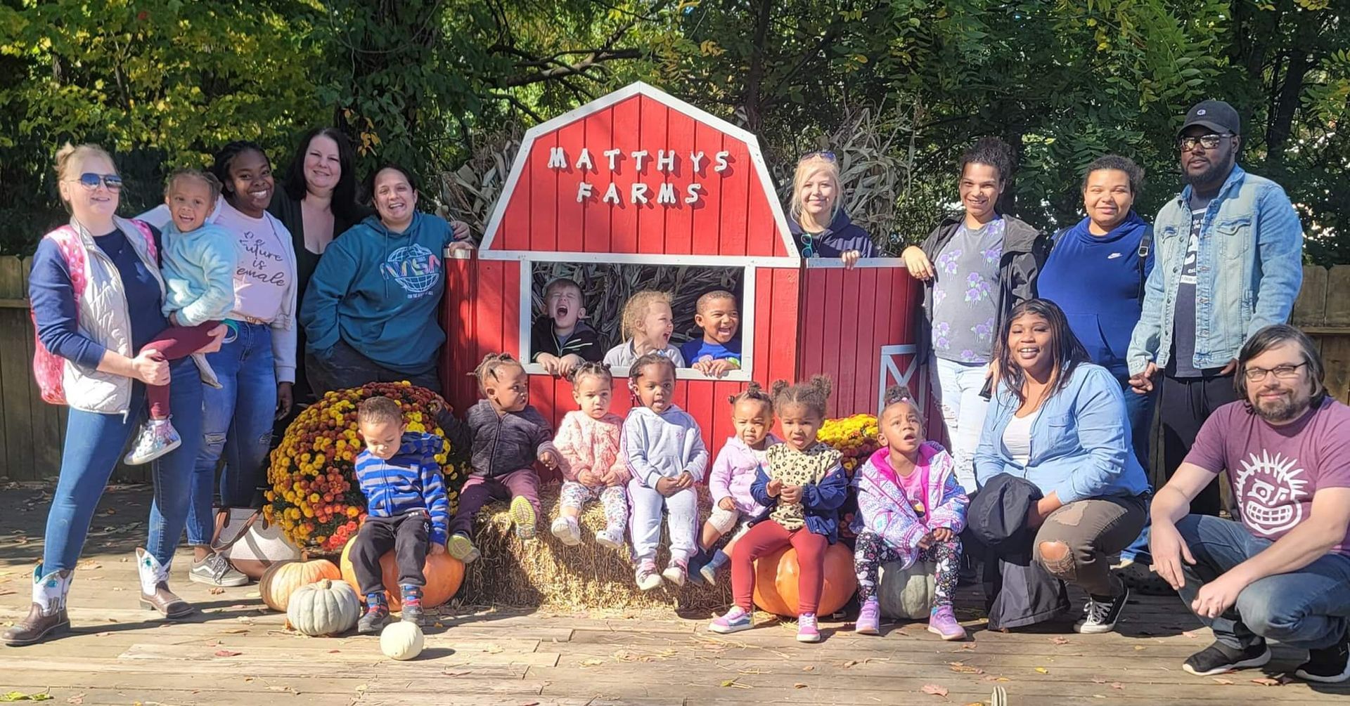 Group posing outdoors in front of a red playhouse, with several children and adults smiling in the sun.