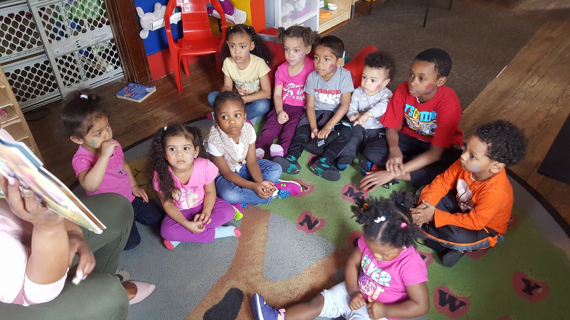 Children sitting in a circle on a classroom floor, listening to a person holding a book.