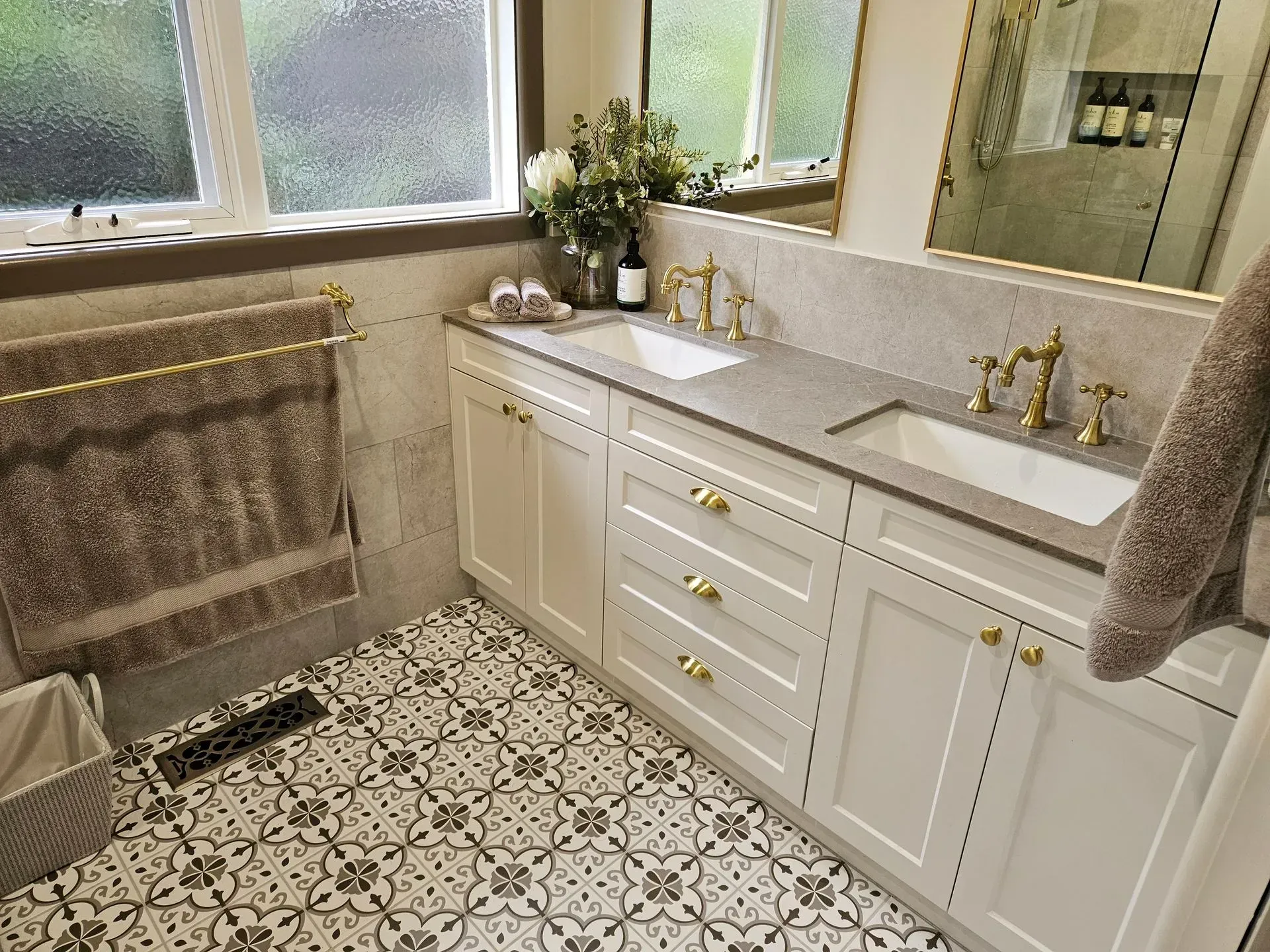 Bright bathroom with white vanity, gold fixtures, patterned floor tile, and towels by a window