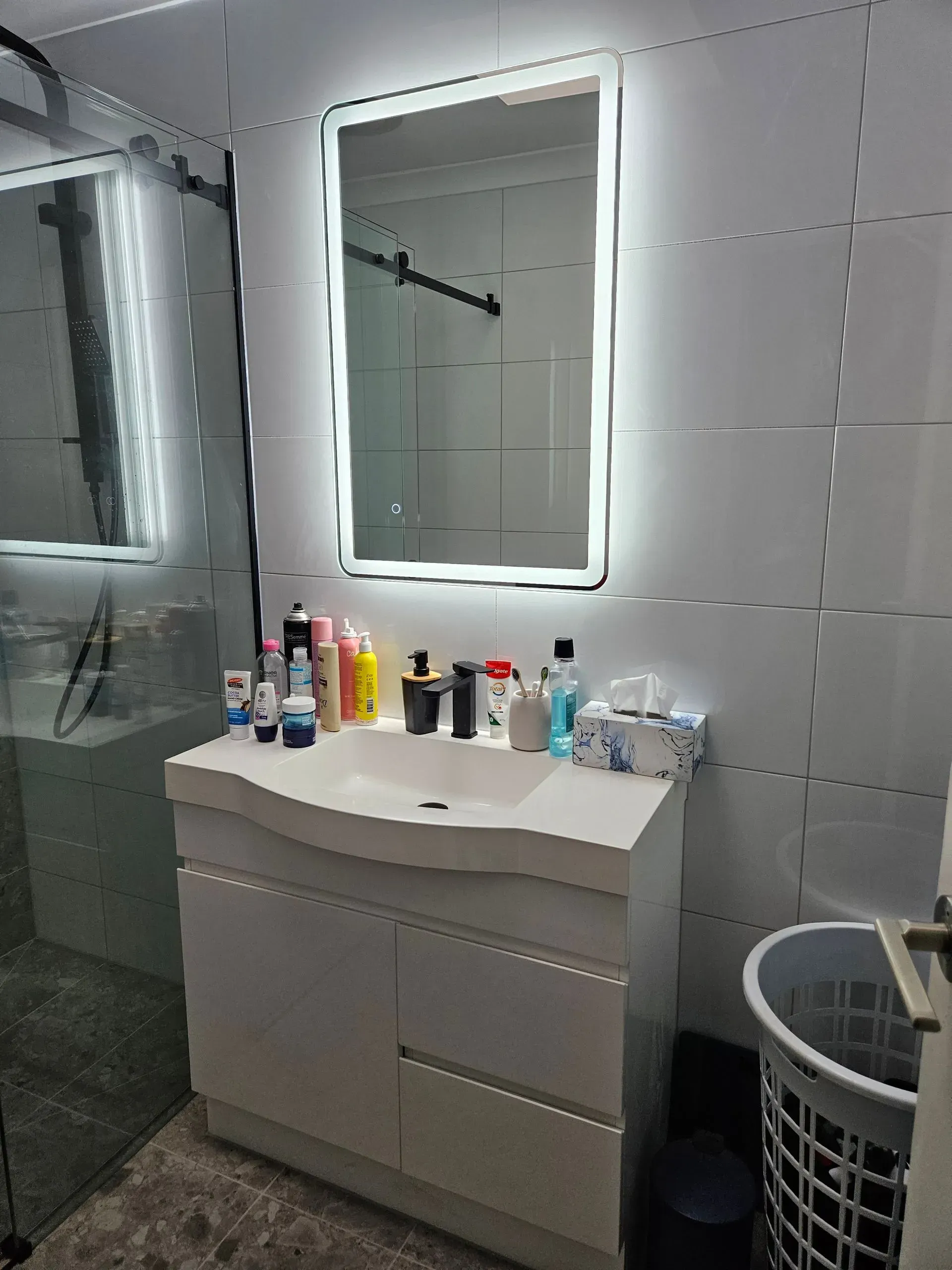 Modern bathroom vanity with lit mirror, sink, toiletries, and laundry basket on the right