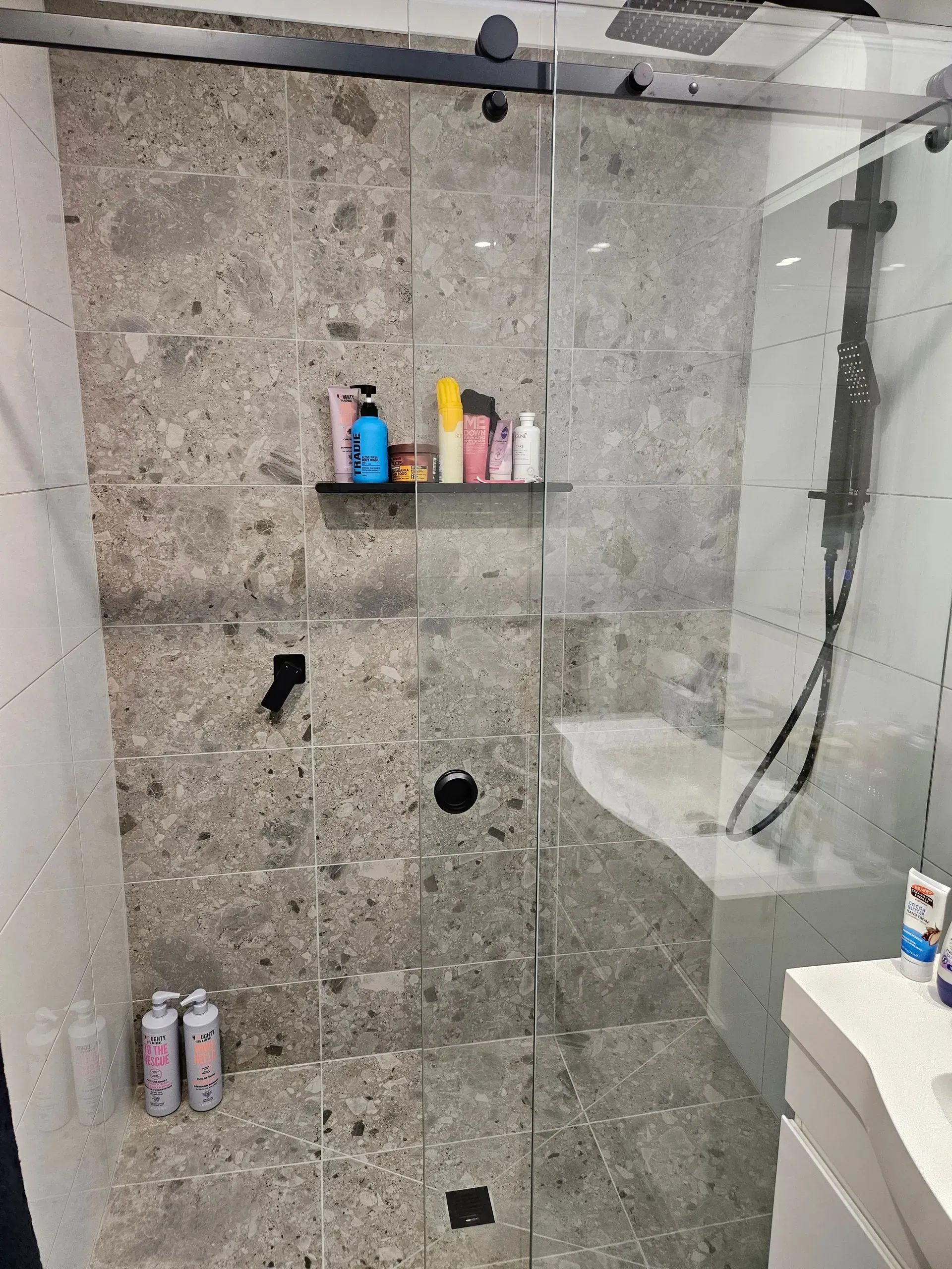Modern shower with gray stone tile, glass door, built-in shelf, and toiletries on a shelf and floor.