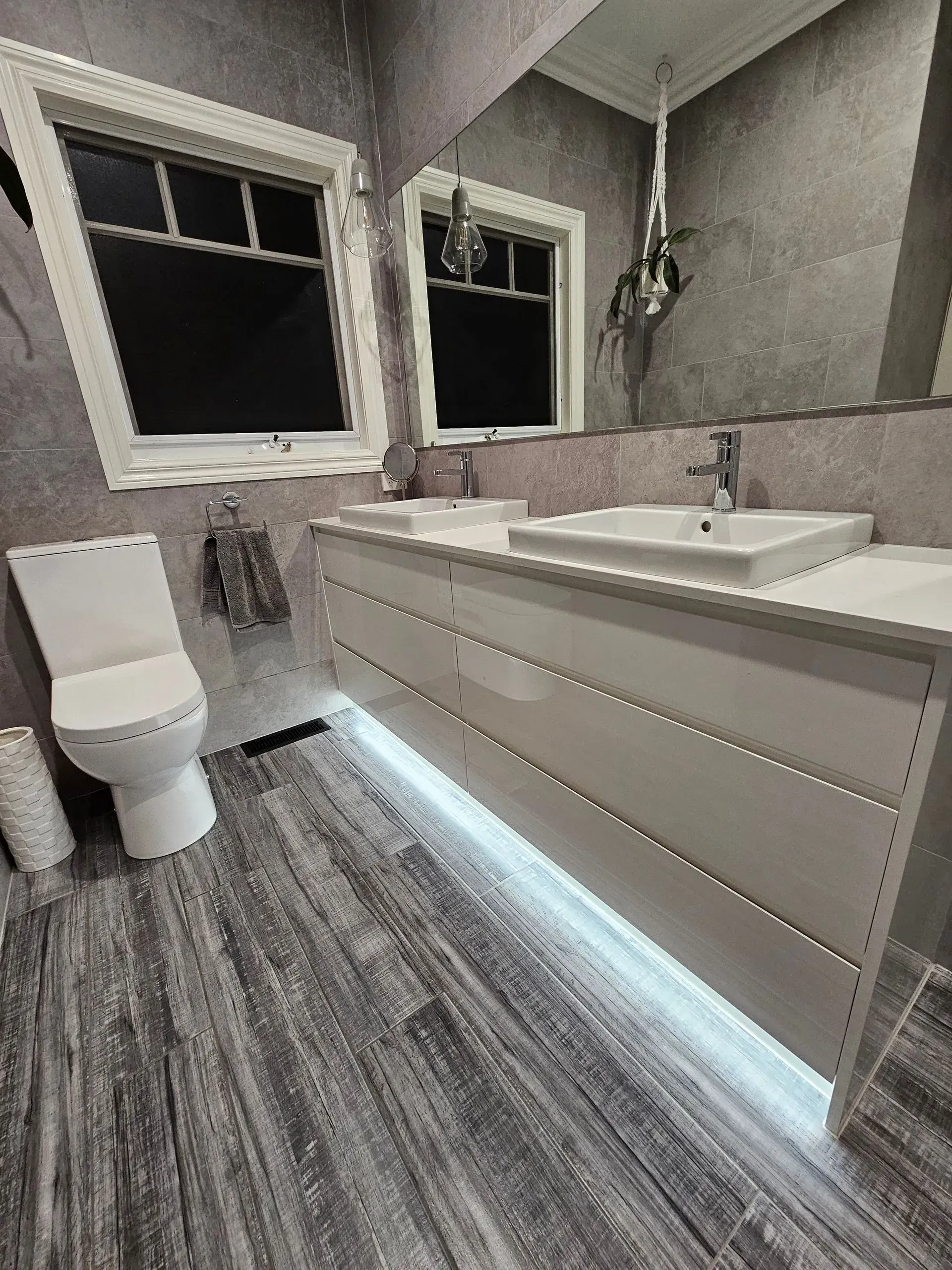 Modern bathroom with double sink vanity, toilet, and under-cabinet lighting near two windows.