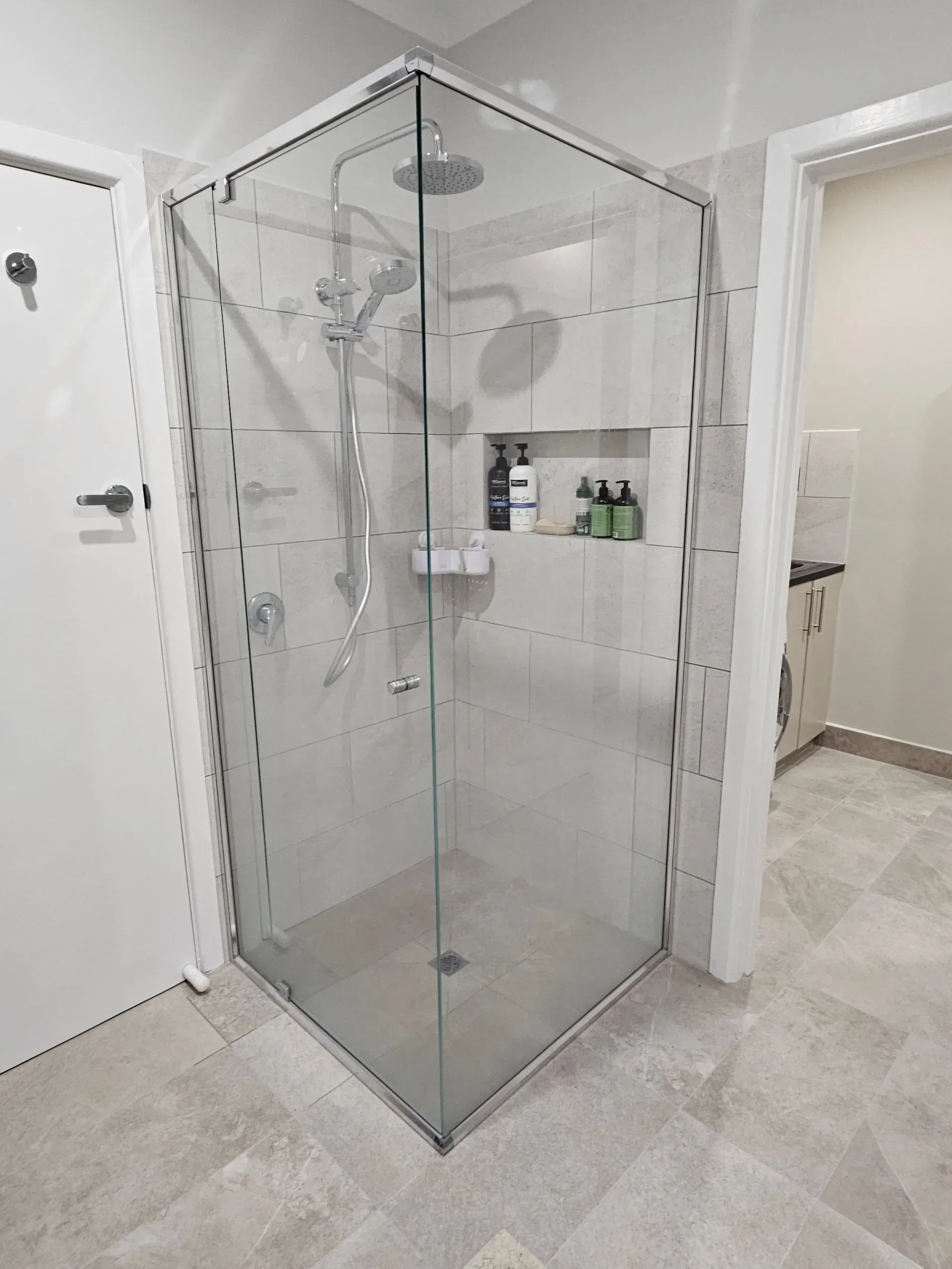 Glass corner shower stall in a light-colored bathroom with tiled walls and floor, seen from the doorway
