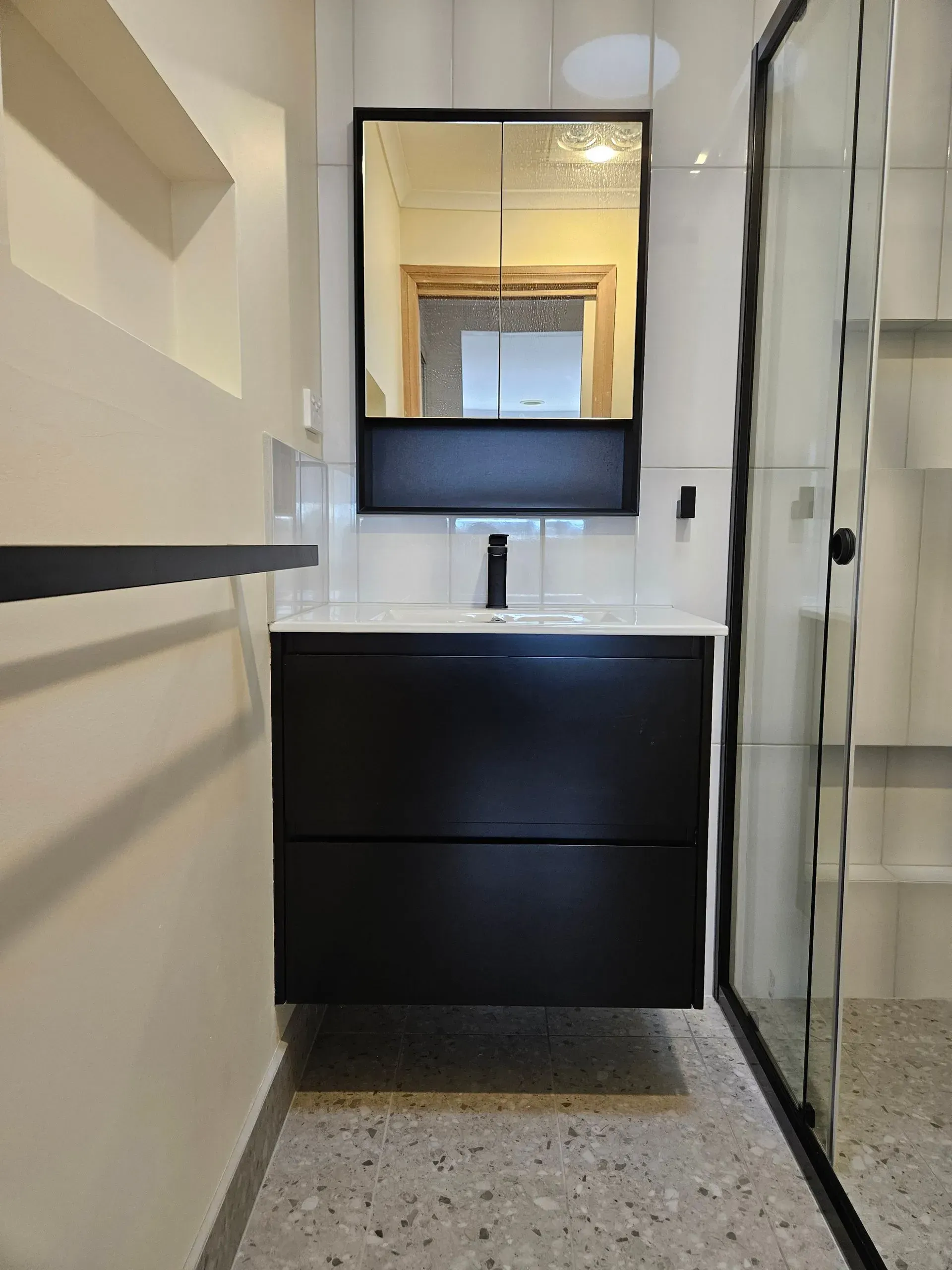 Narrow modern bathroom with black vanity, mirror, white walls, and glass shower door.