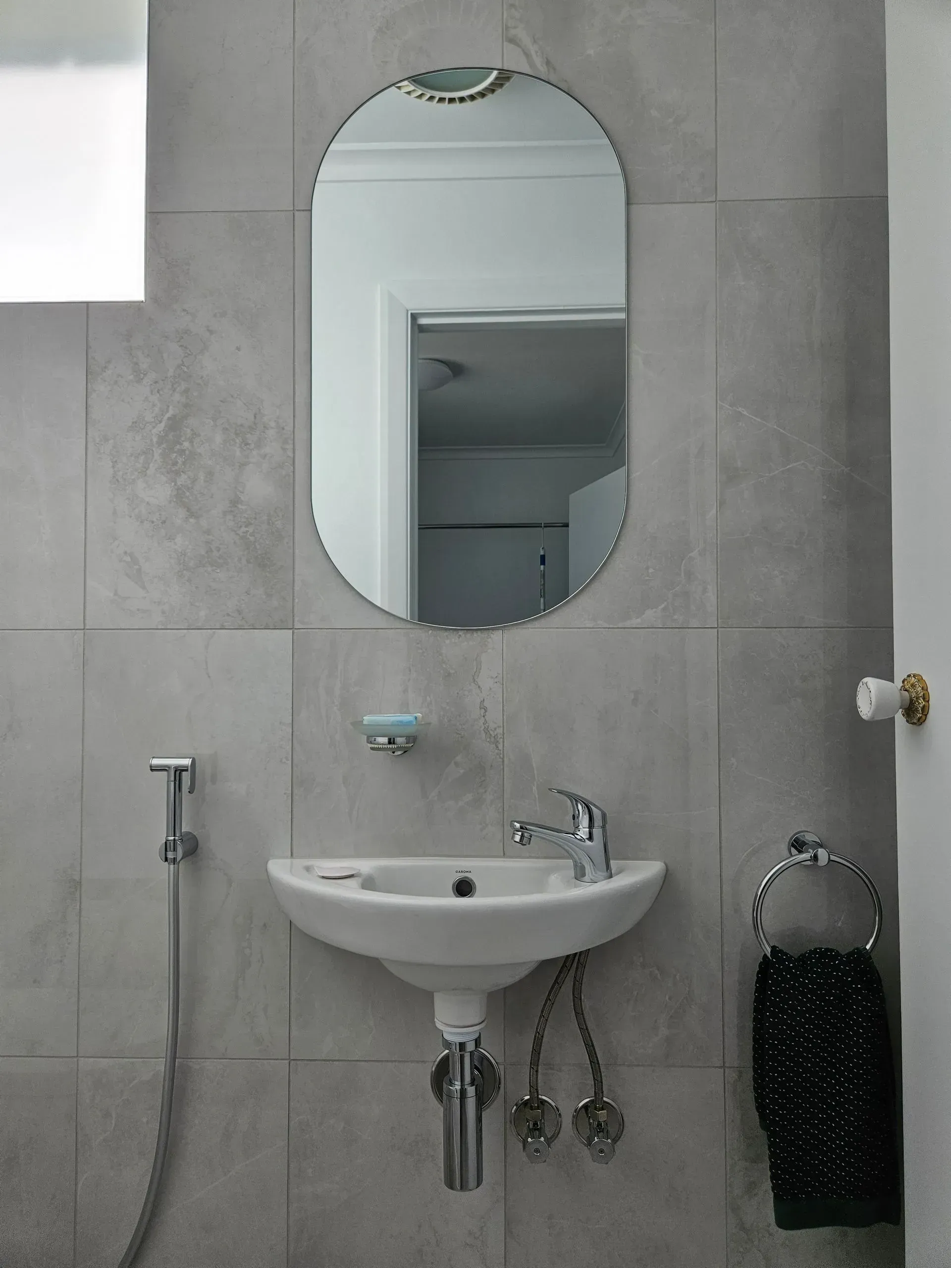 Gray tiled bathroom with oval mirror, wall-mounted sink, and towel rack.