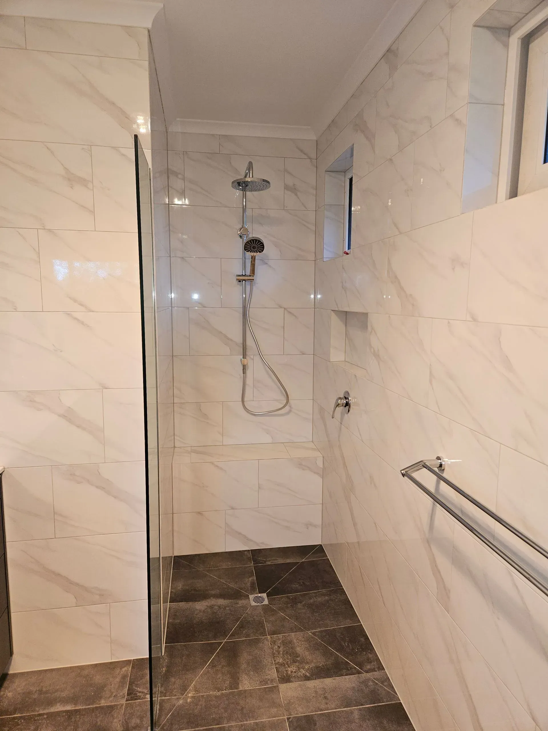 Small tiled shower room with a wall-mounted showerhead, grab bar, and dark wet-looking floor.