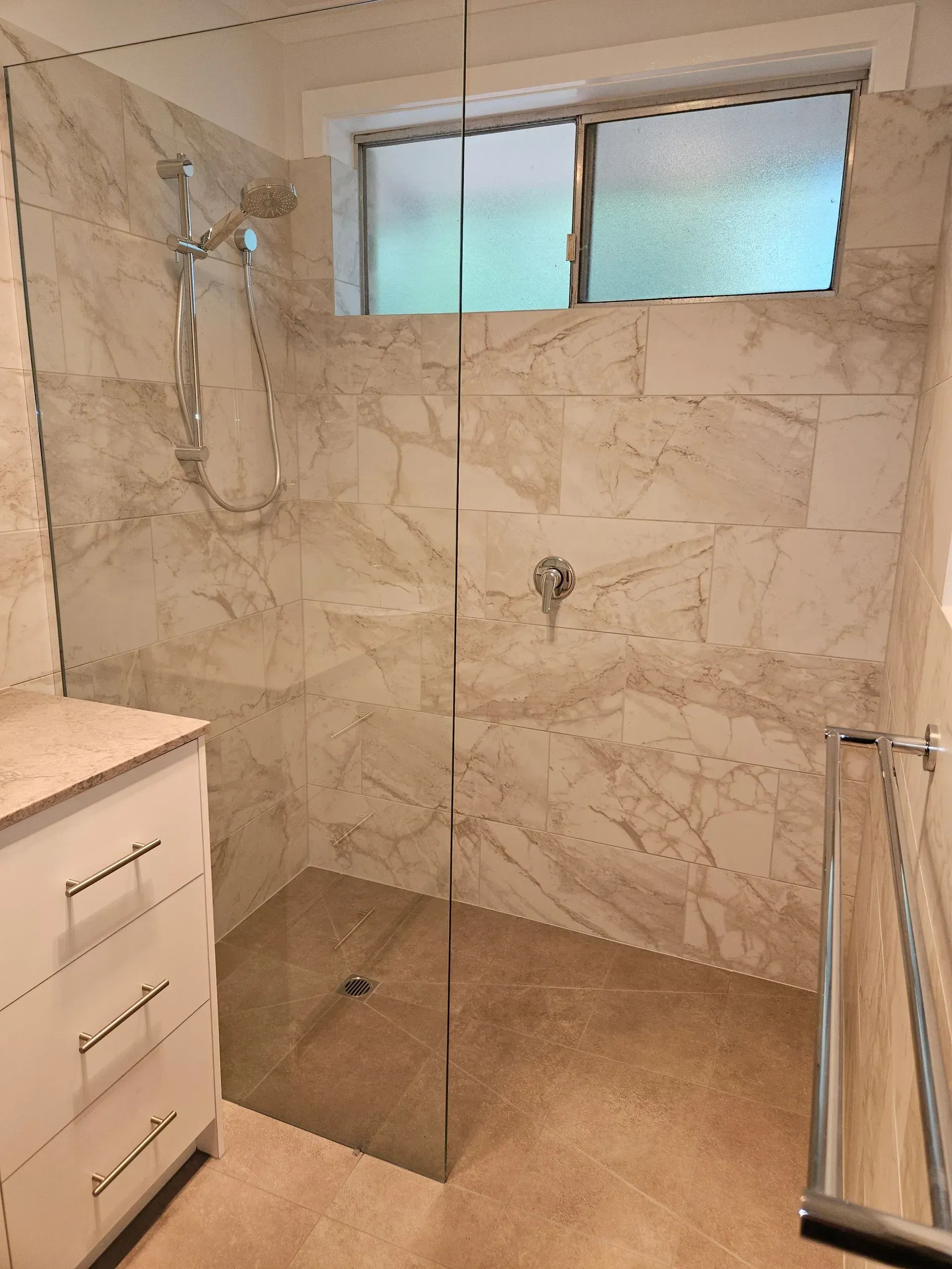 Modern bathroom shower with marble tile, glass enclosure, window, and white vanity cabinet