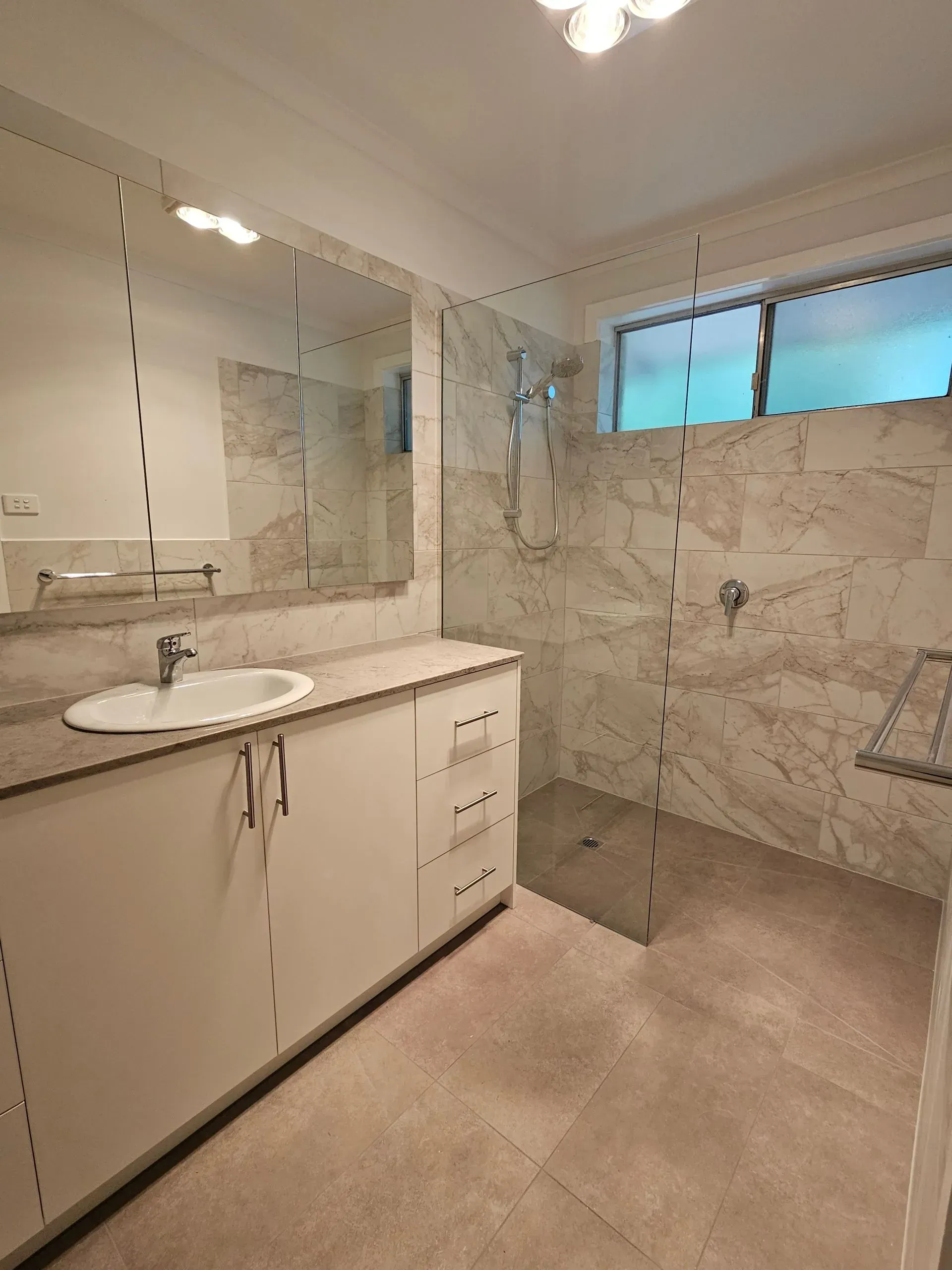 Modern bathroom with white vanity, large mirror, glass shower, and marble tile walls