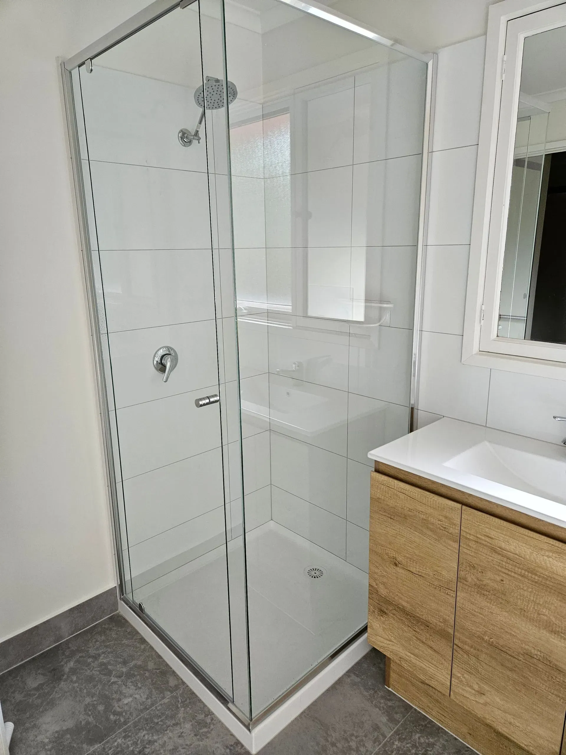 Glass shower enclosure in a modern bathroom beside a wooden vanity and mirror.