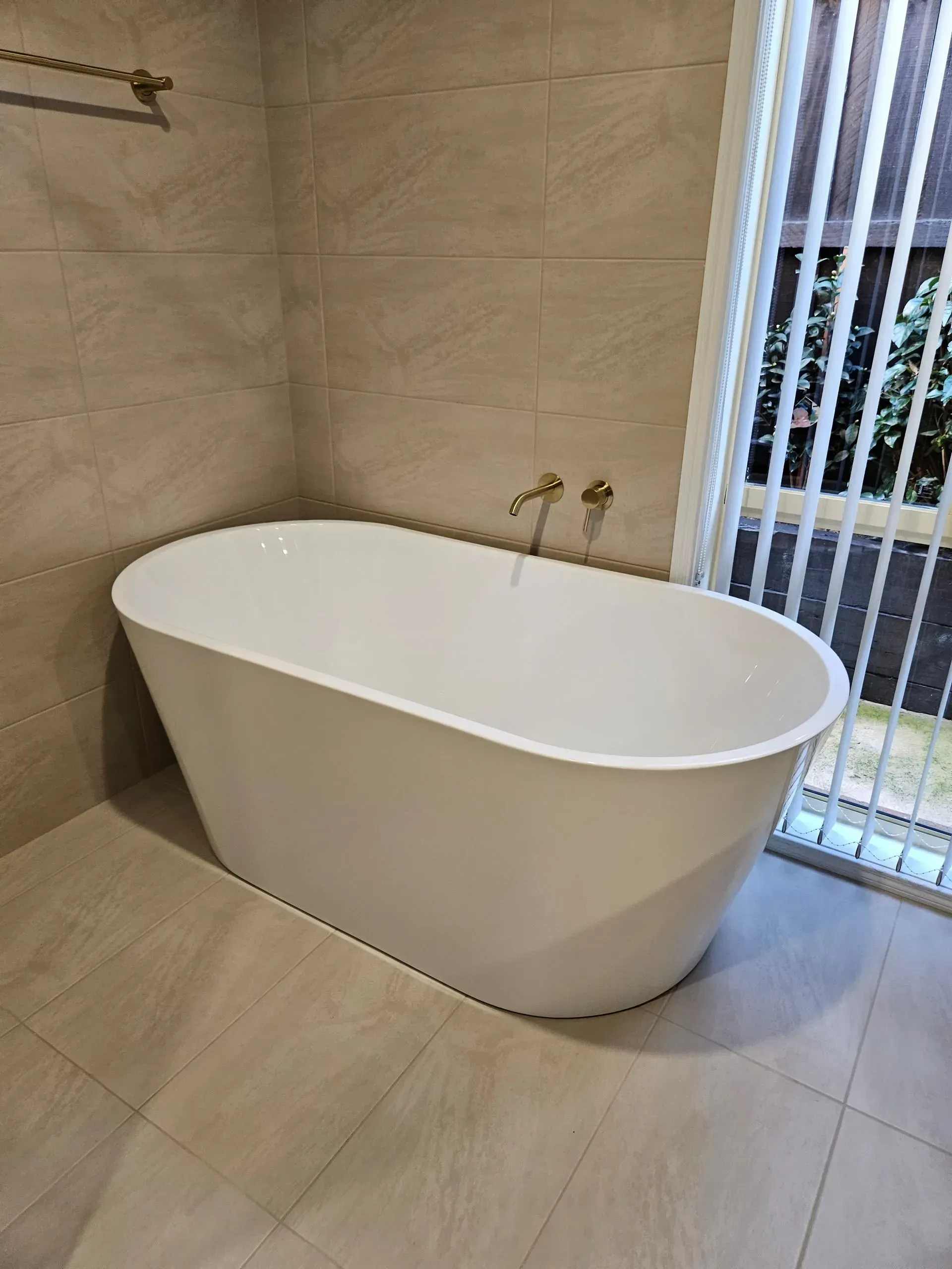 Modern white freestanding bathtub in a beige-tiled bathroom beside a window with vertical blinds
