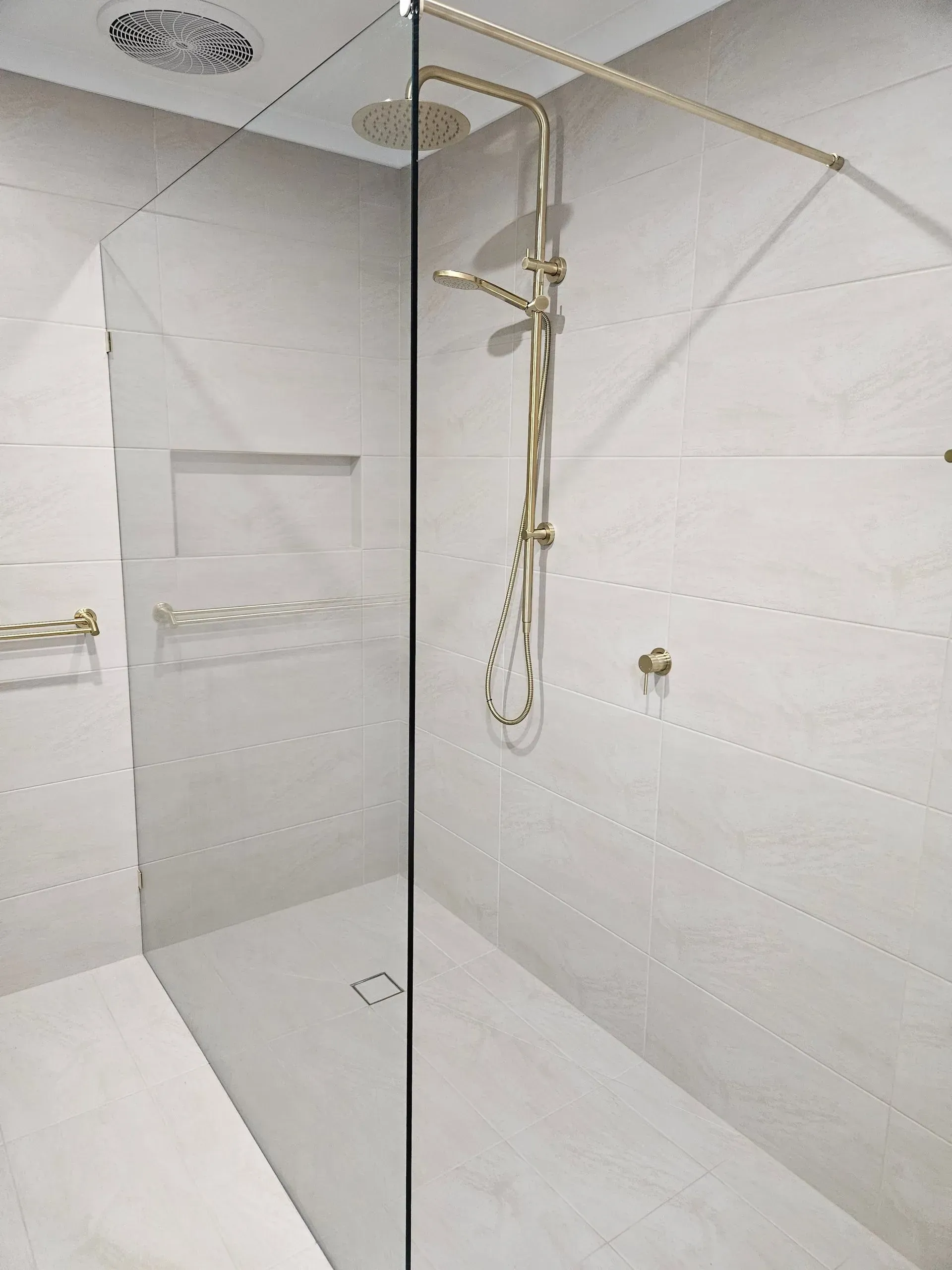 Modern white tiled shower with glass partition, rainfall showerhead, handheld sprayer, and built-in shelf