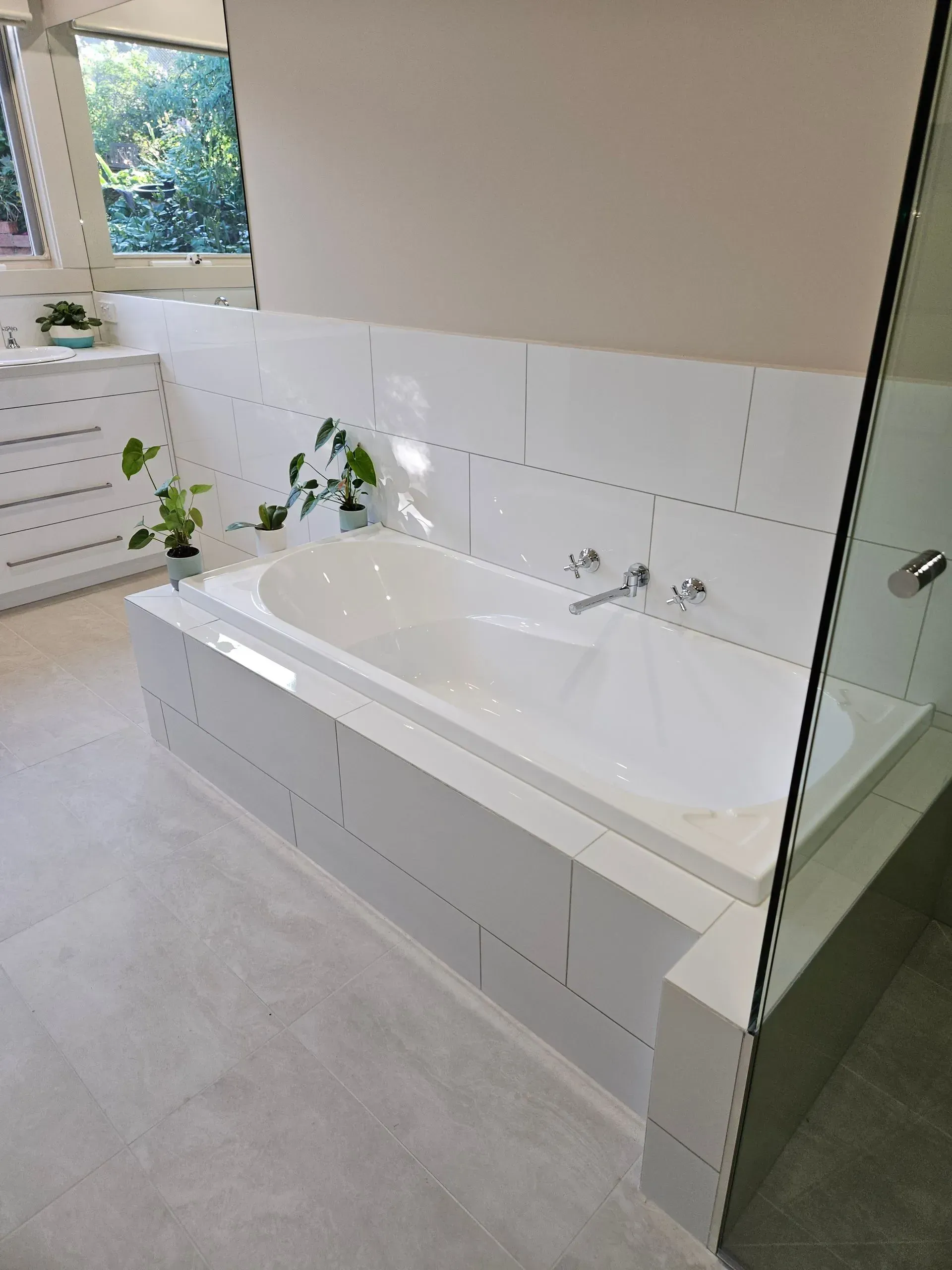 Modern white bathroom with a built-in bathtub, tiled walls, and a window with plants.