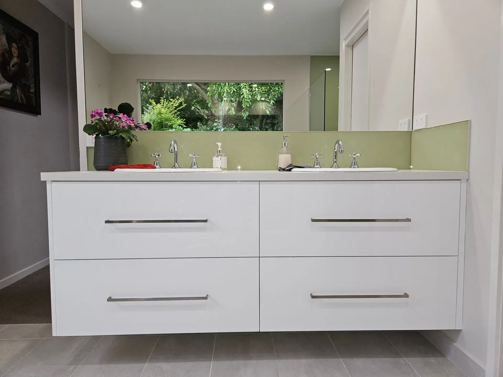Modern bathroom vanity with double sink, white cabinets, mirrors, and window with flowers.