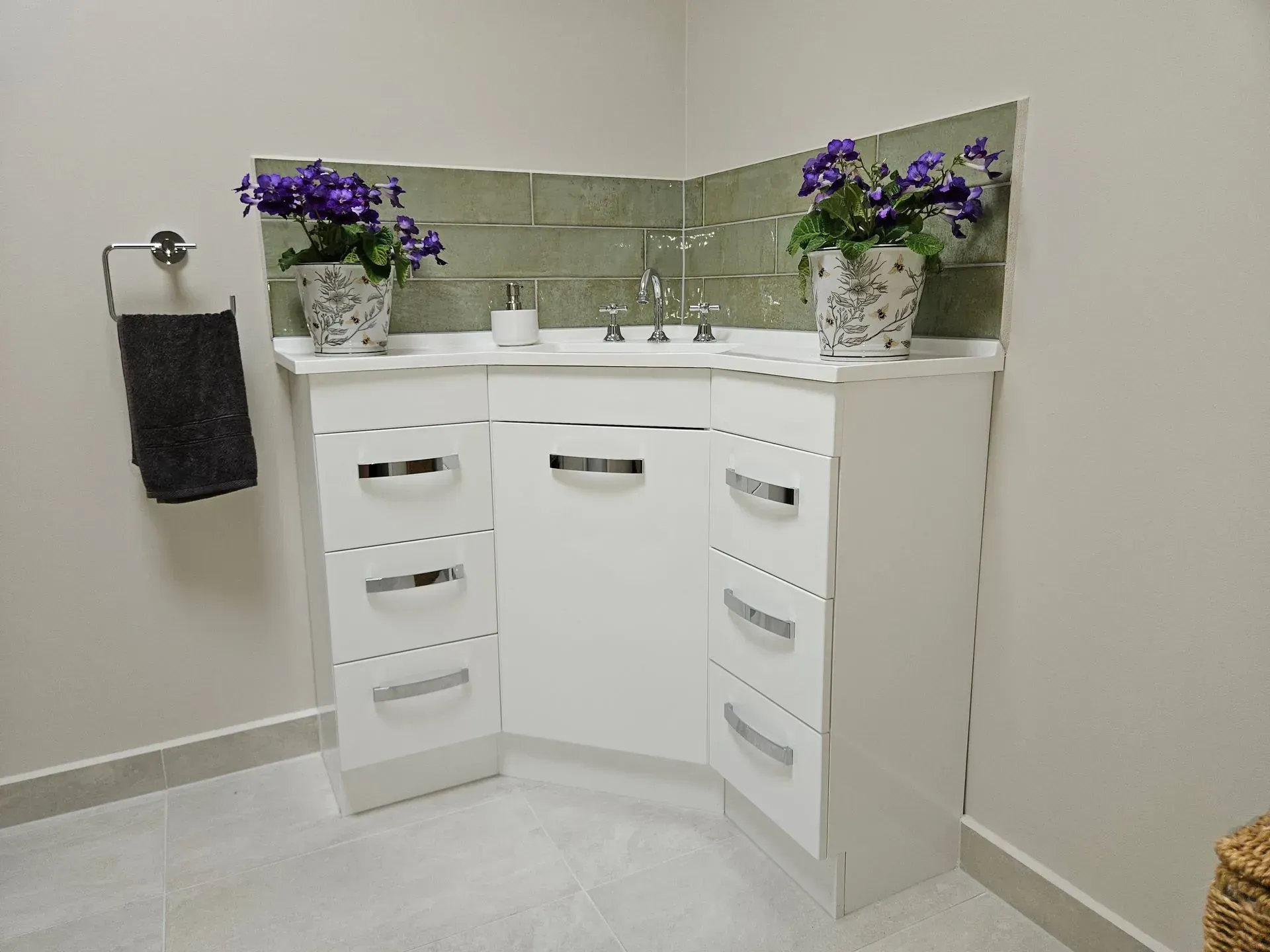 White bathroom vanity with corner sink, mirror, drawers, and purple flowers on the counter.