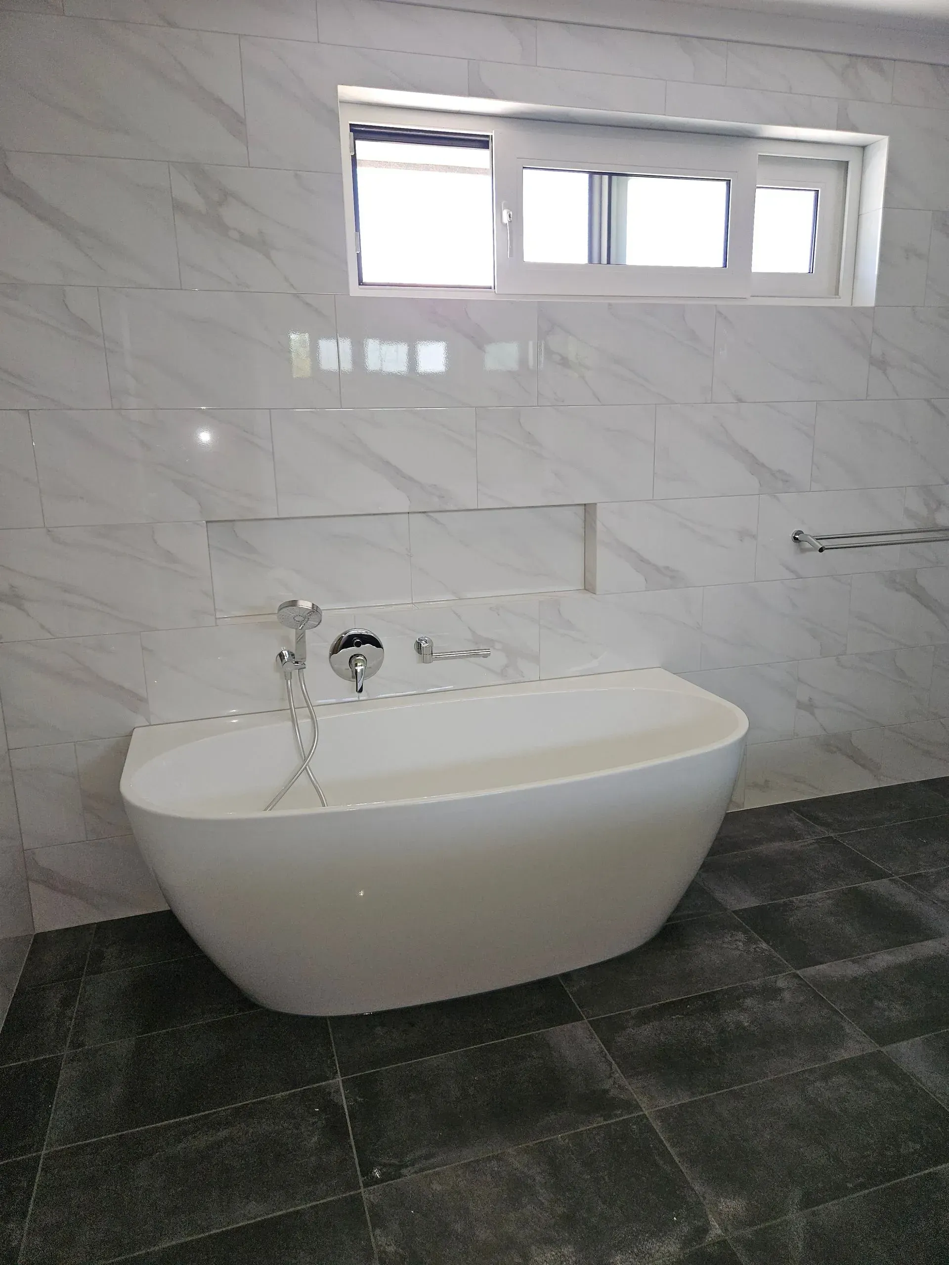 Modern bathroom with a white wall-mounted bathtub, marble tiles, and a small window above.