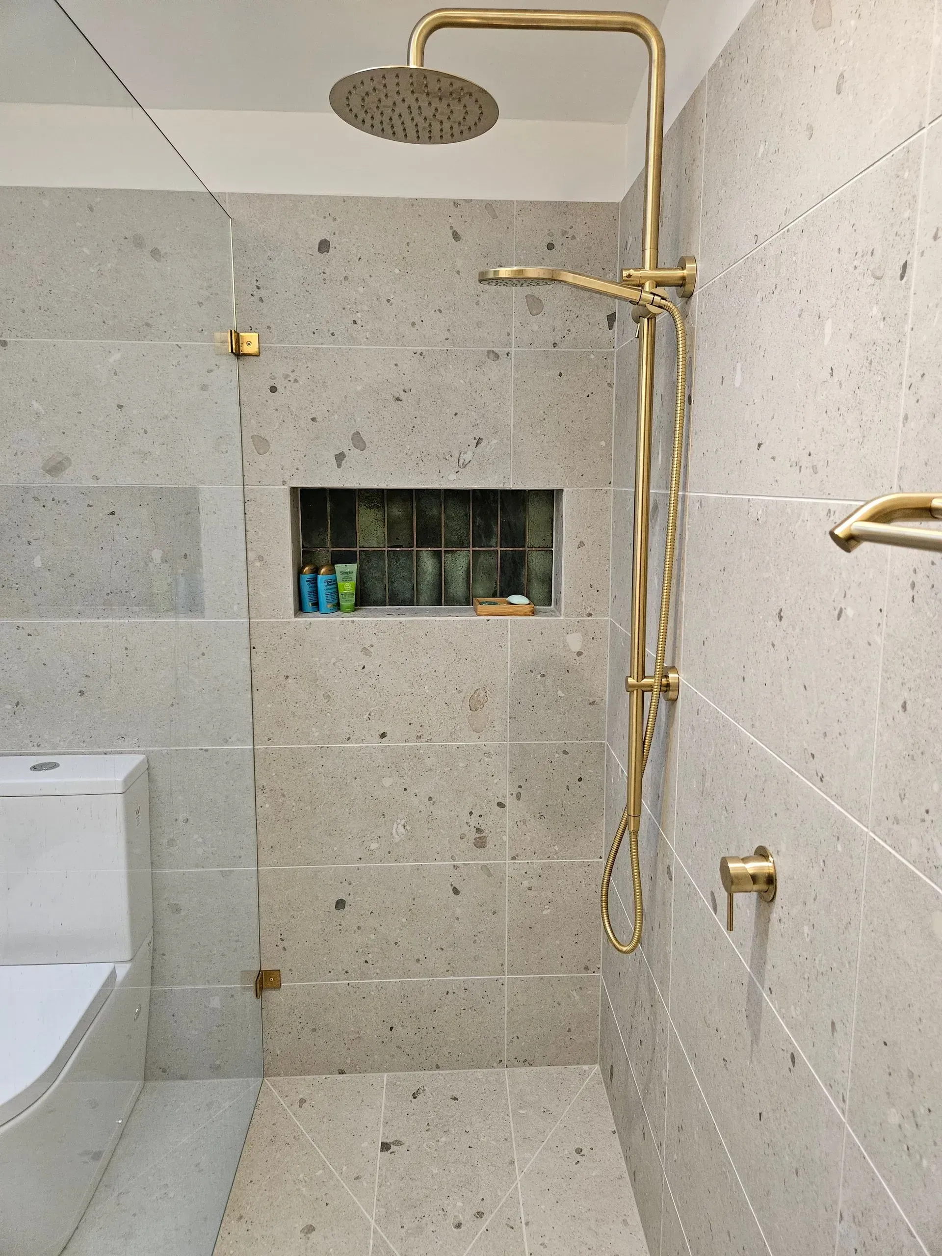 Modern shower with beige tile, gold fixtures, and built-in wall niche with toiletries