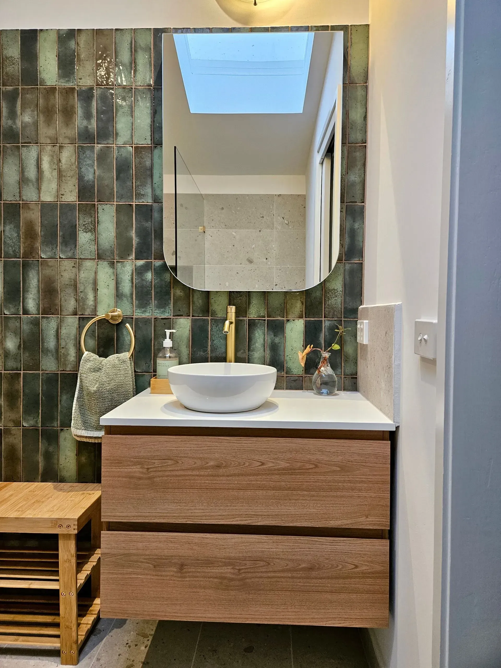 Small bathroom vanity with round sink, wood drawers, green tile wall, mirror, skylight, and stool.