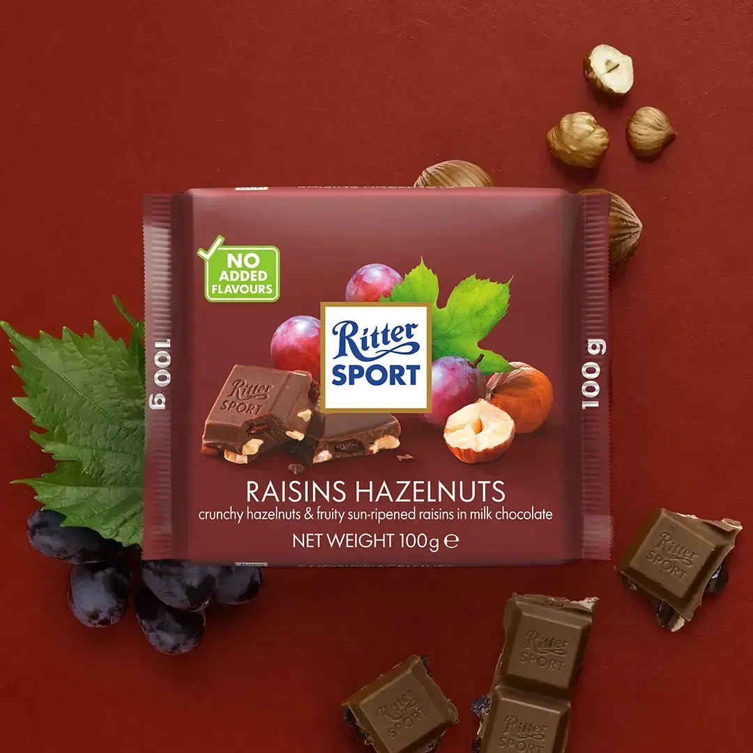 Ritter Sport chocolate bar with raisins and hazelnuts, on a red background with grapes and nuts.