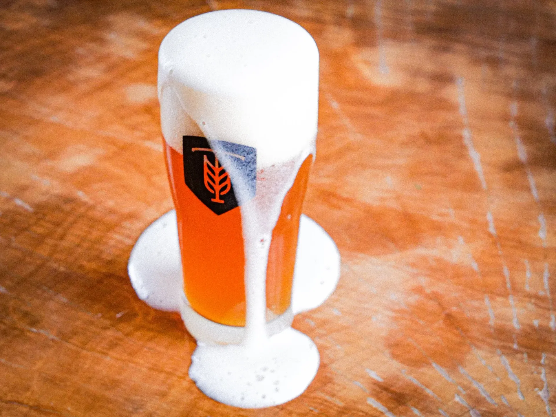 Glass of beer overflowing onto a wooden surface. Foam pooling around the glass.