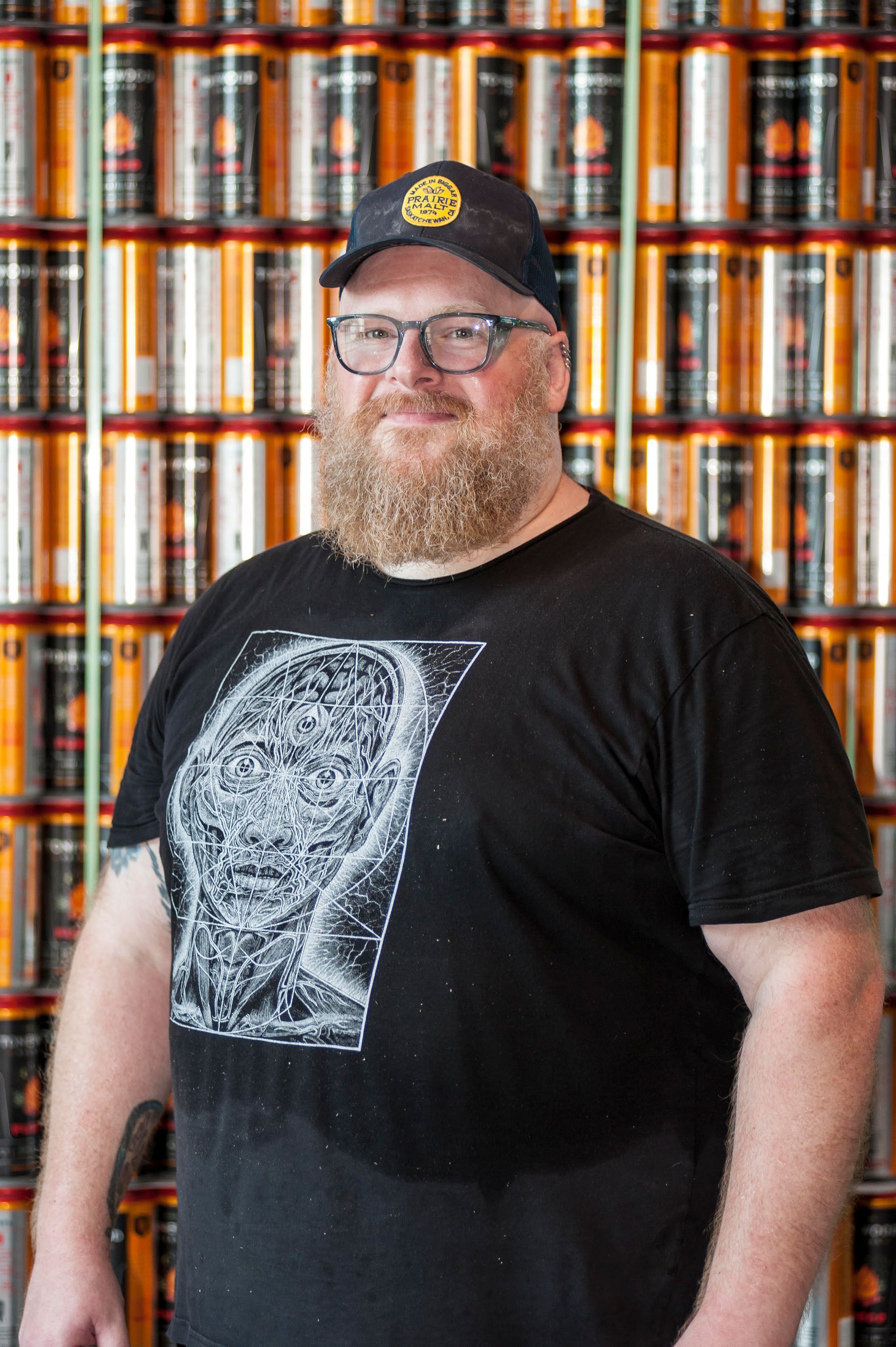 Smiling person in a black graphic T-shirt and cap standing before a wall of stacked cans