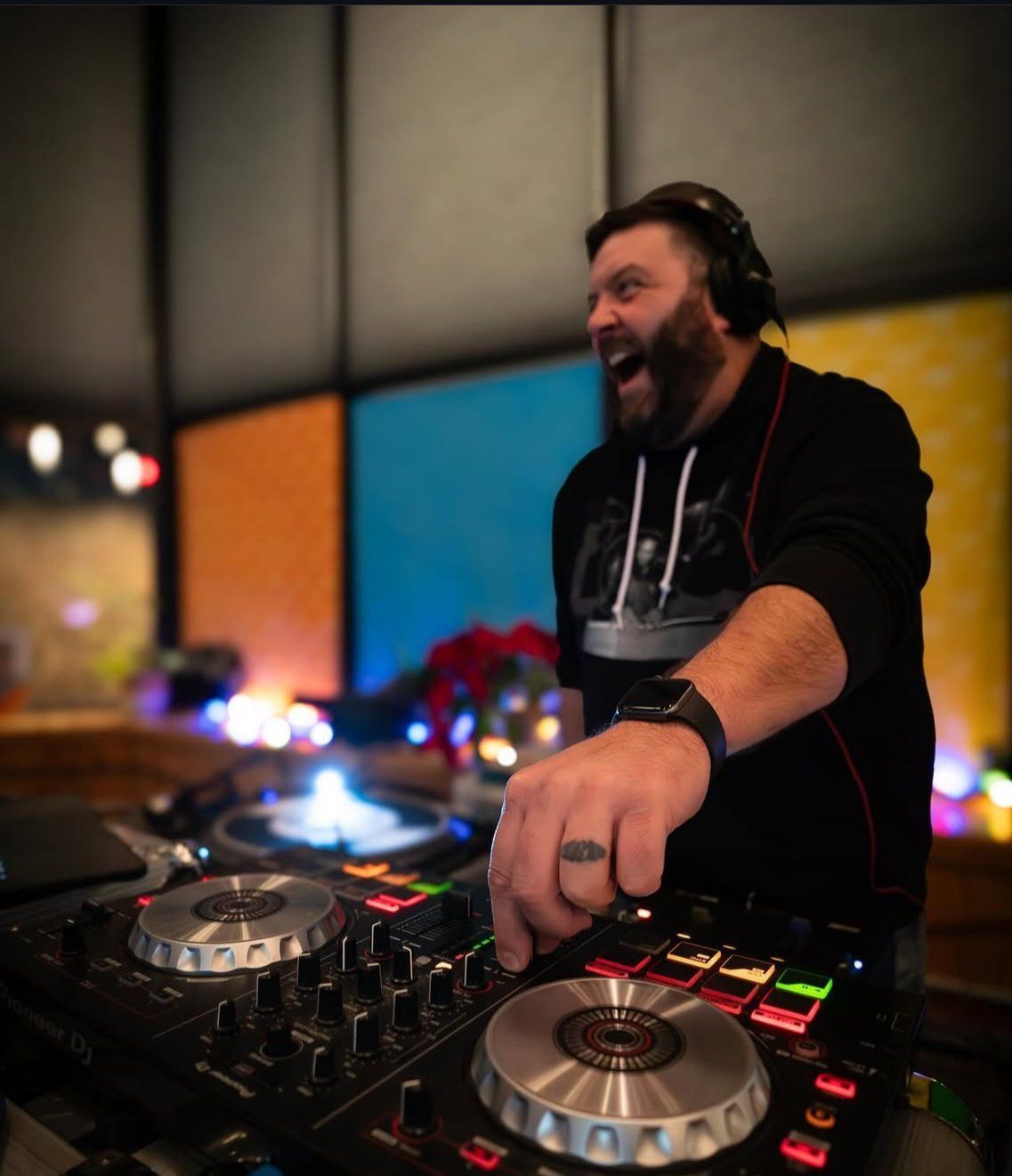 DJ smiling, mixing music. Wearing headphones, black hoodie. Colorful background.