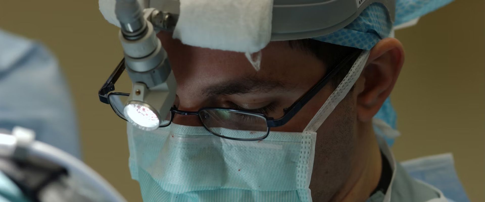 A surgeon wearing a mask and glasses is operating on a patient.