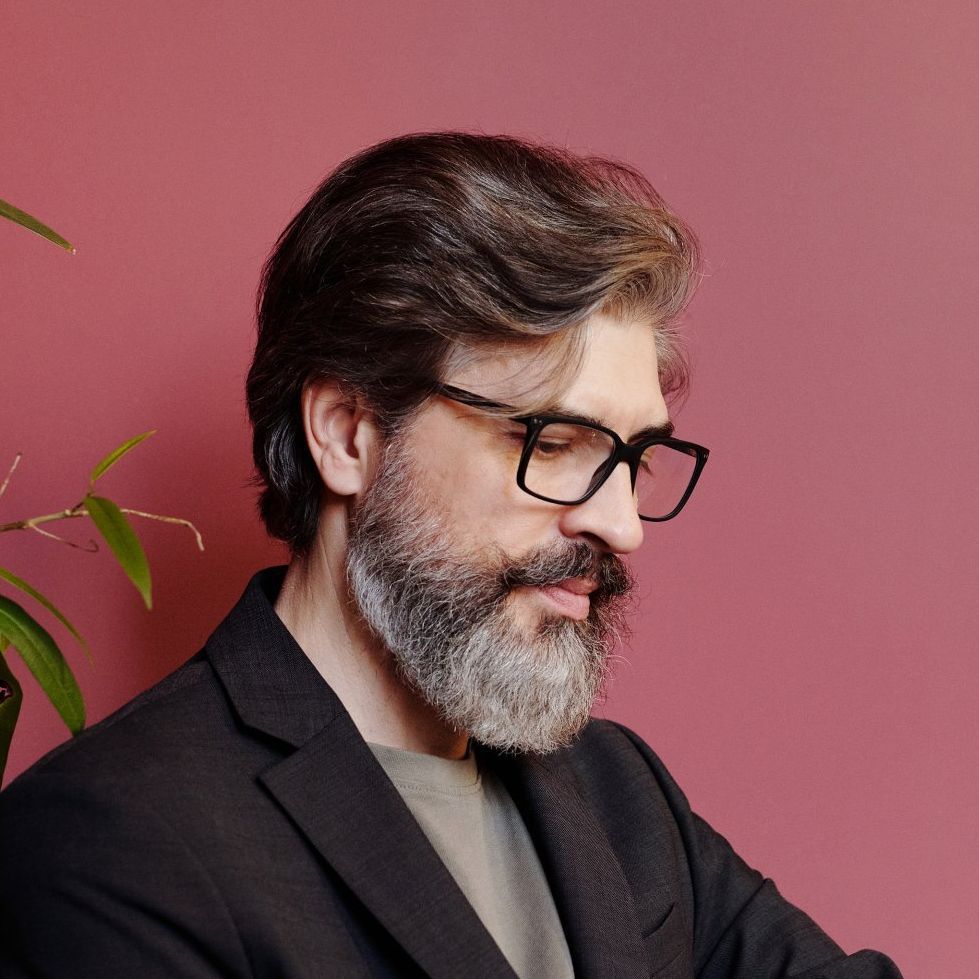 A man with a beard and glasses is sitting in front of a pink wall.