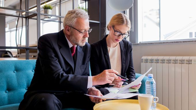 Man in suit points at papers, woman looks on, seated on blue couch near window.