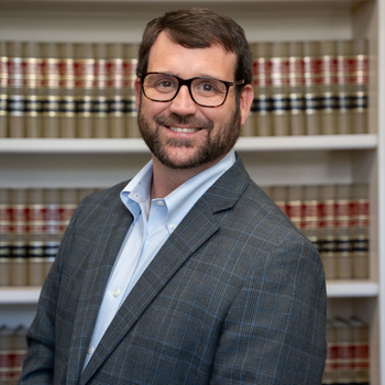 Man in glasses and suit smiles in front of a bookshelf.