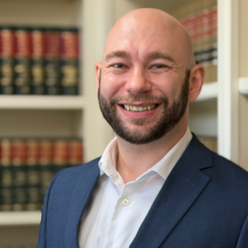 Man in glasses and suit smiles in front of a bookshelf.