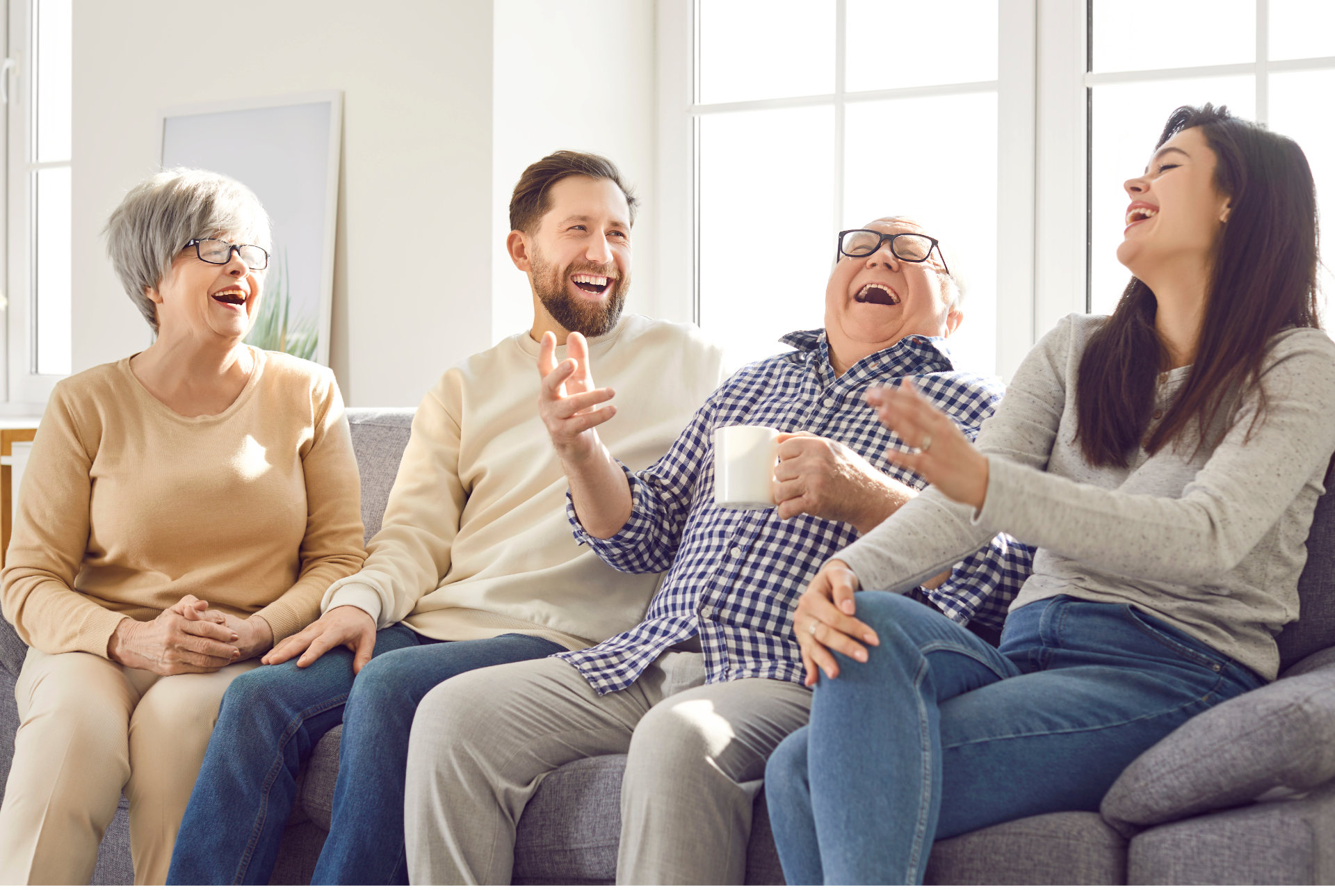 Four people laughing together on a couch indoors: two older women, and a man. Sunlight streams in.
