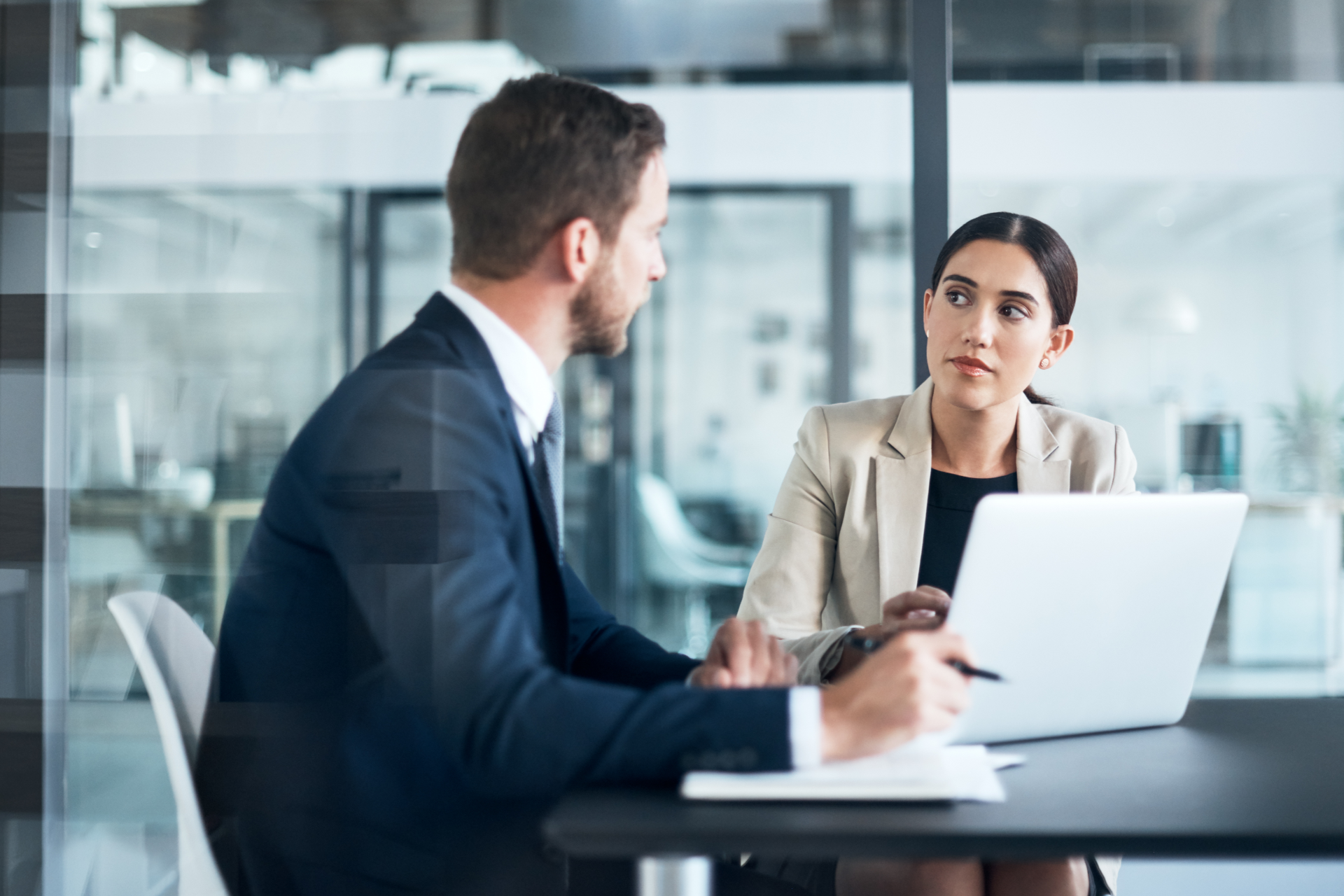 Man and woman in business attire discuss around a laptop in a modern office.