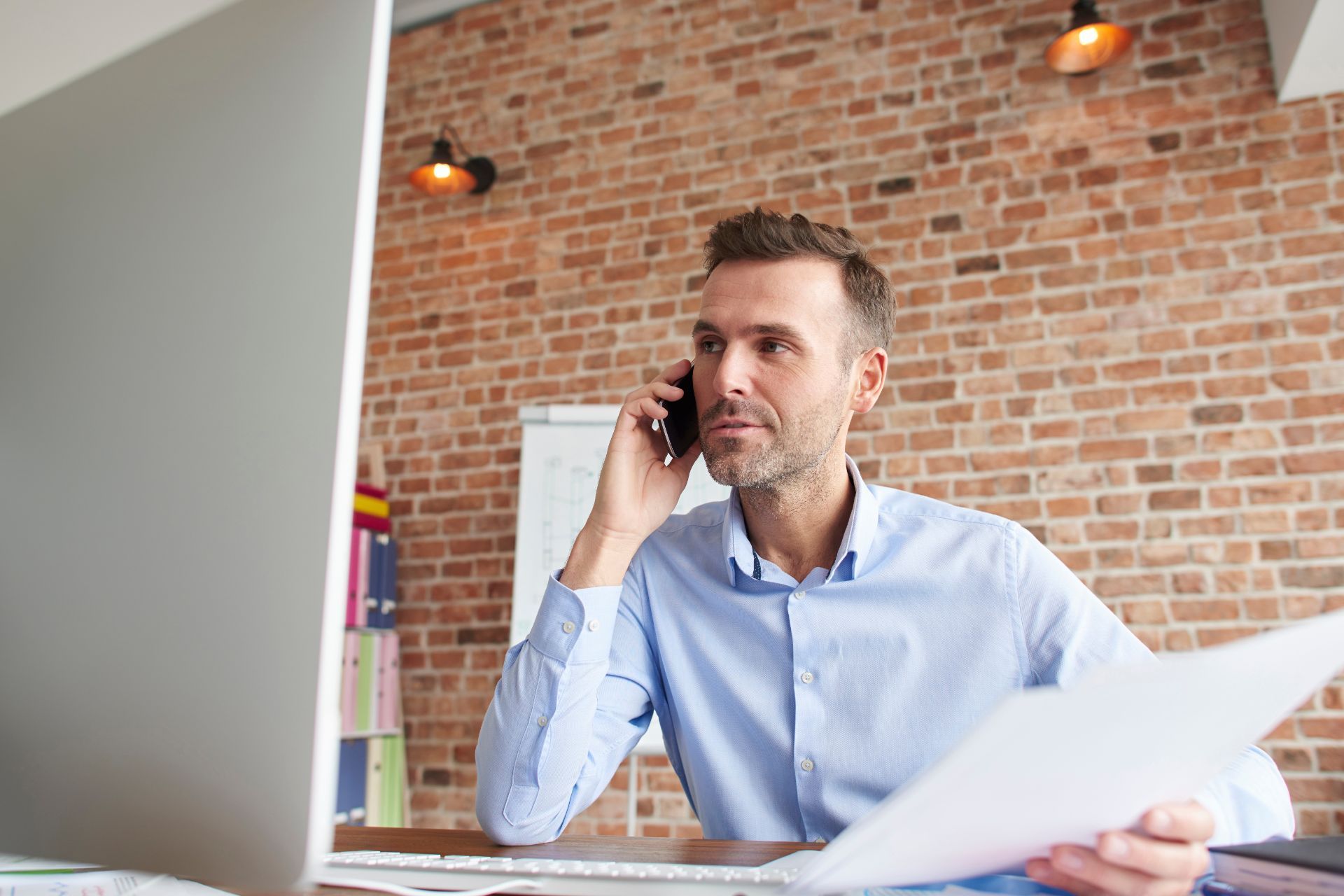 Man on phone in office, holding paper, looking at computer, brick wall background.