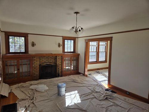 Living room with a brick fireplace, windows, bare floor, and drop cloths during renovation