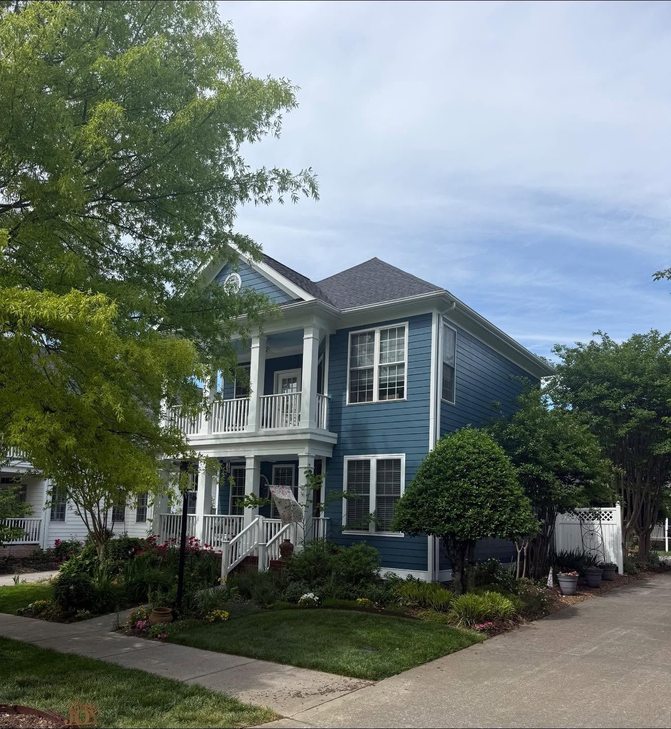 Blue two-story house with white trim, front porch, and a tree-lined driveway on a sunny day