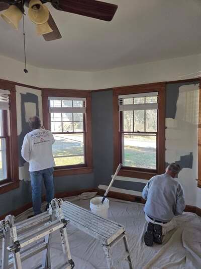 Two workers painting a sunroom with large windows and a ceiling fan, surrounded by ladders and tools.