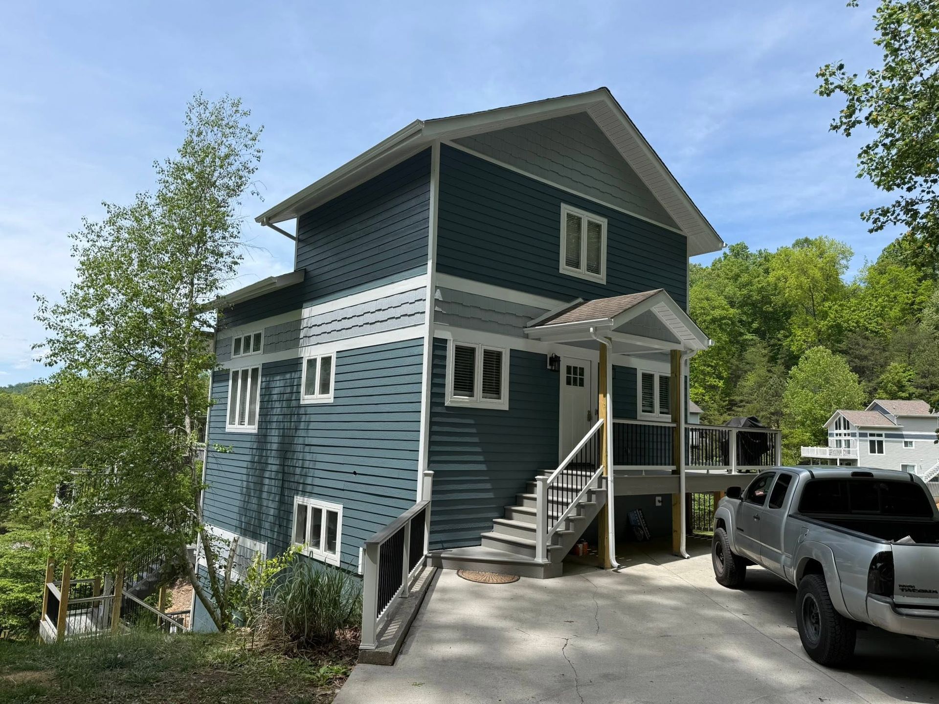 Blue two-story house with white trim, front porch, and parked truck in a wooded driveway