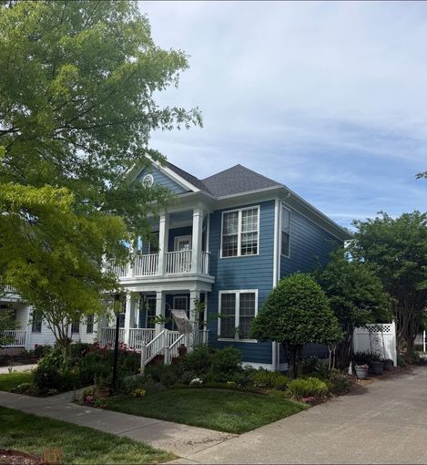 Blue two-story house with white trim, porch, and front garden beside a driveway under a partly cloudy sky