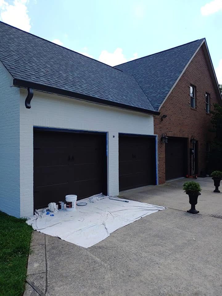 White brick house with two black garage doors, brick wing, and a driveway with paint supplies on a tarp
