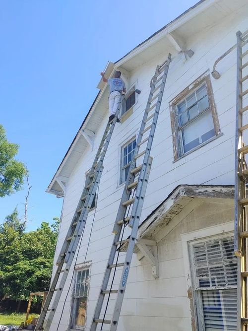 Worker on a tall ladder repairing the roof and upper wall of a white house