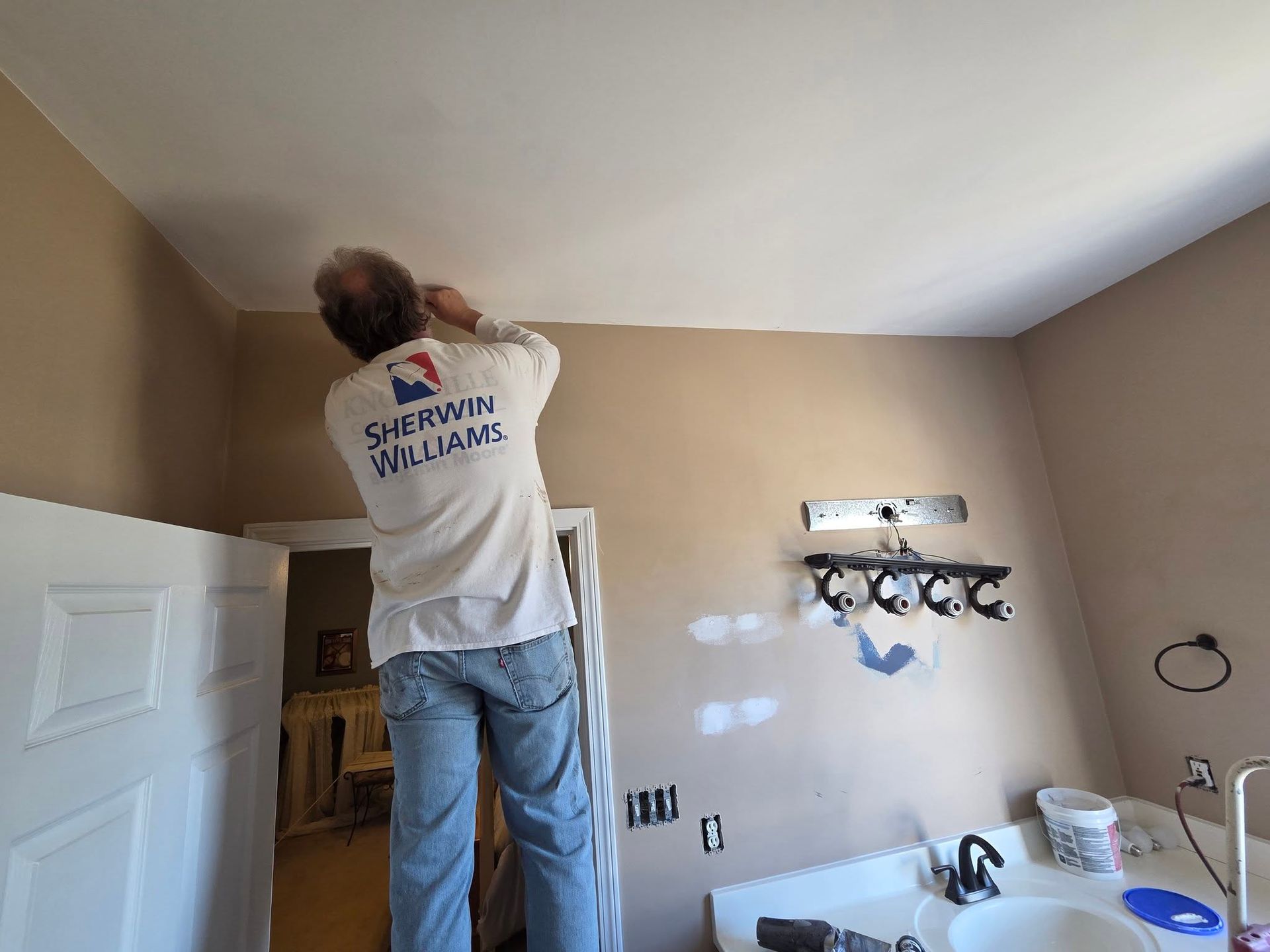 Person painting a beige bathroom ceiling while standing on a step ladder, with vanity lights and sink visible.