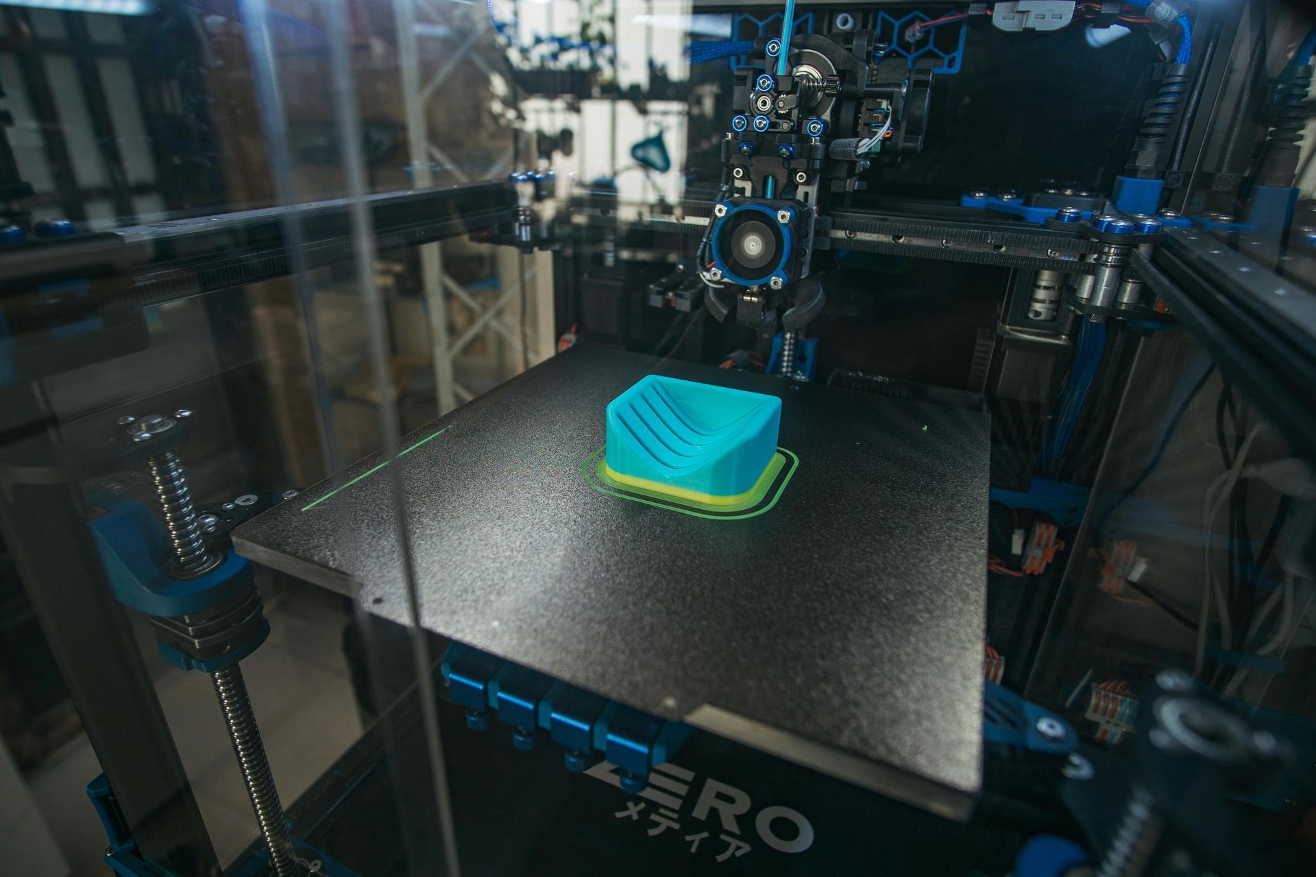 3D printer printing a turquoise object on a black build plate inside an enclosed frame