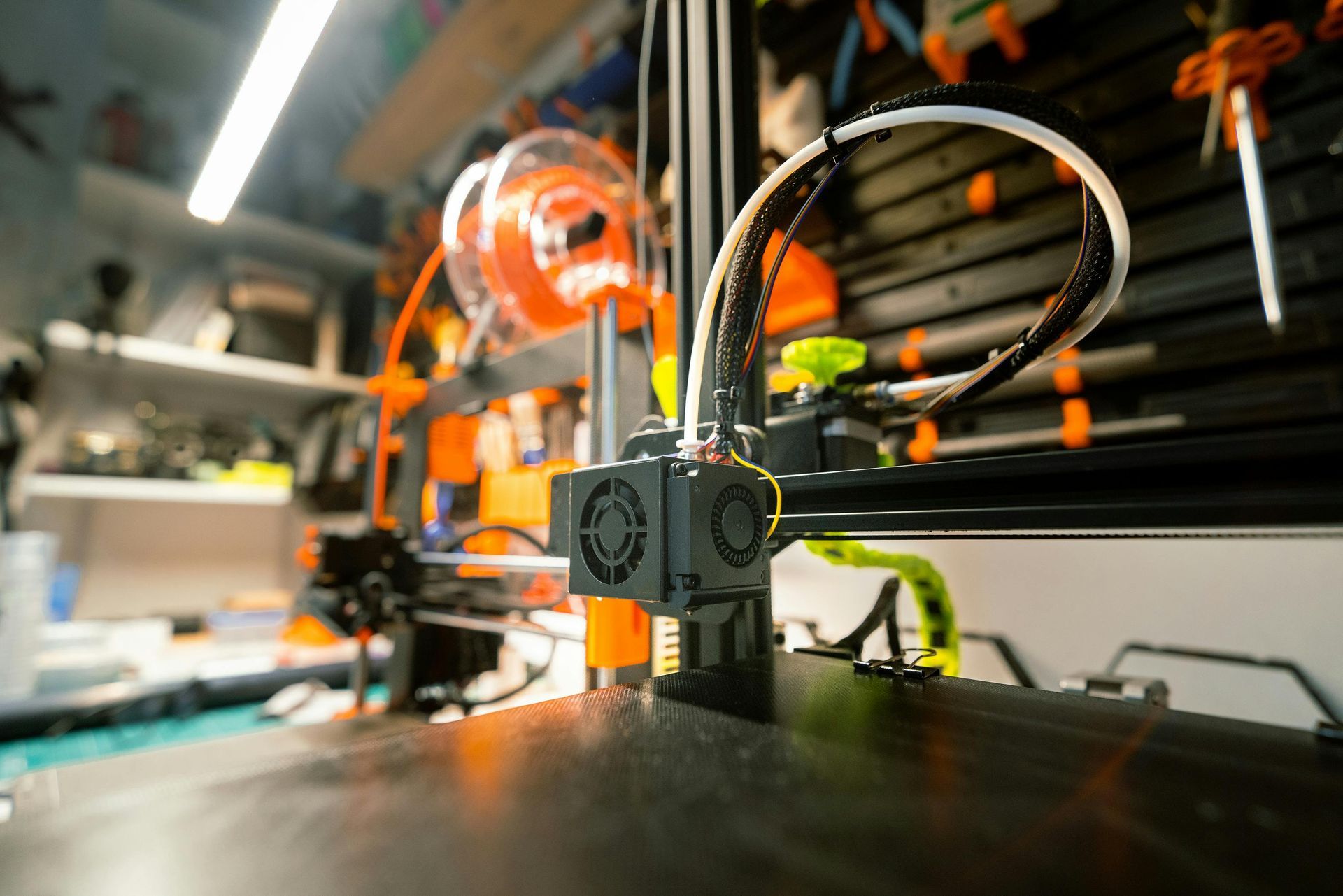 3D printer nozzle over a print bed in a workshop with orange filament and blurred tools in the background
