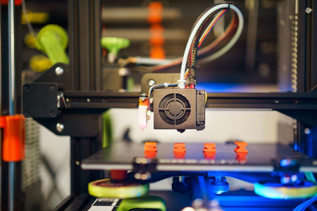 3D printer actively printing a model on a build plate, with a cooling fan and filament visible