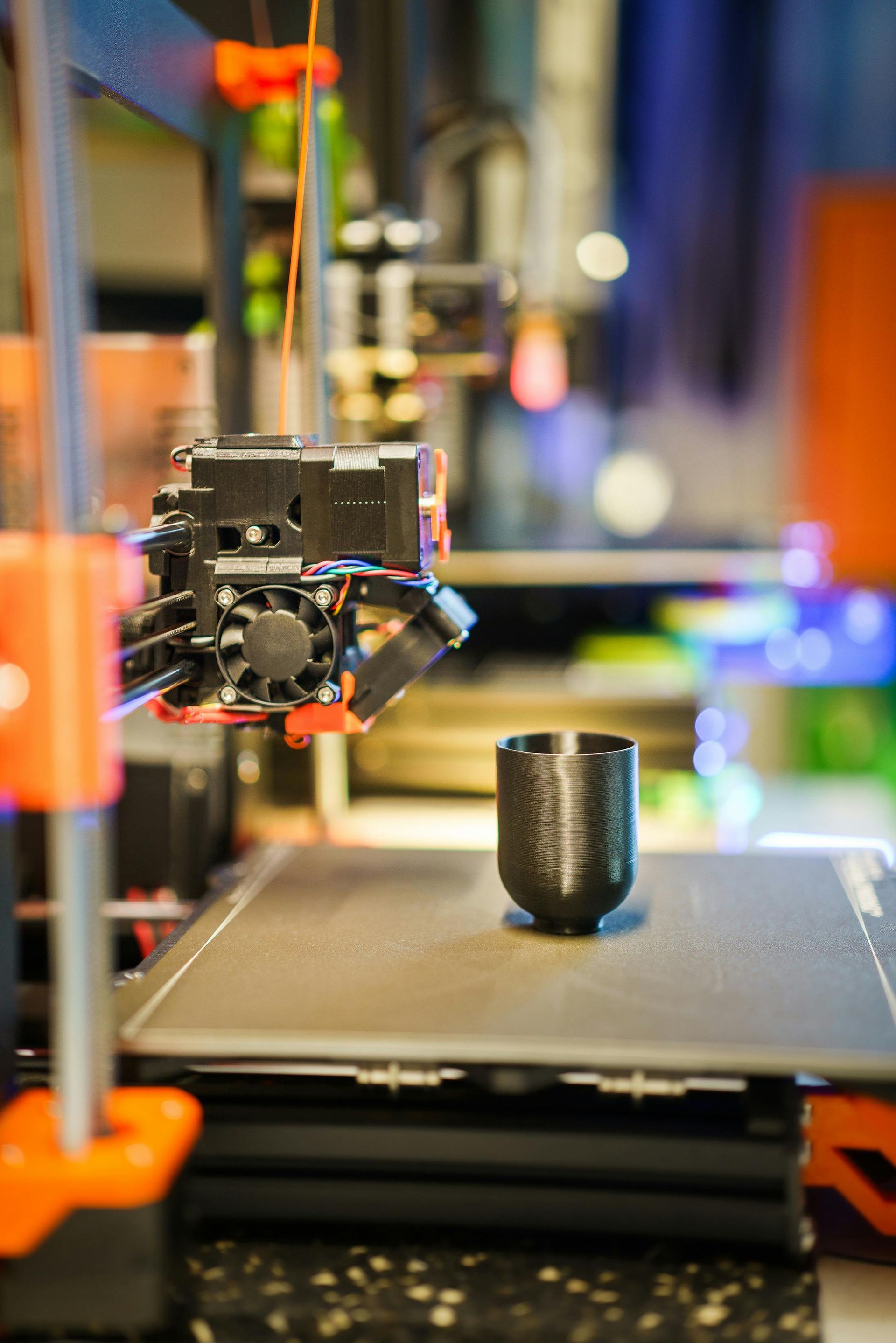 3D printer printing a black cup on a build plate with a blurred workshop background