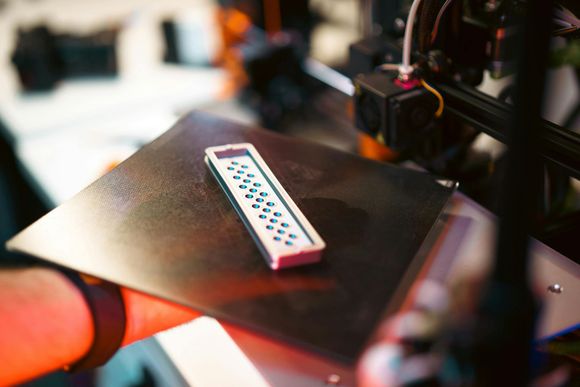 Hand holding a black tray with a white remote control on top in a blurred workshop setting