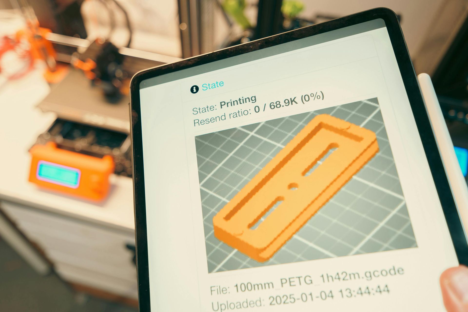 Tablet showing 3D printer control screen with an orange printed part preview in a workshop