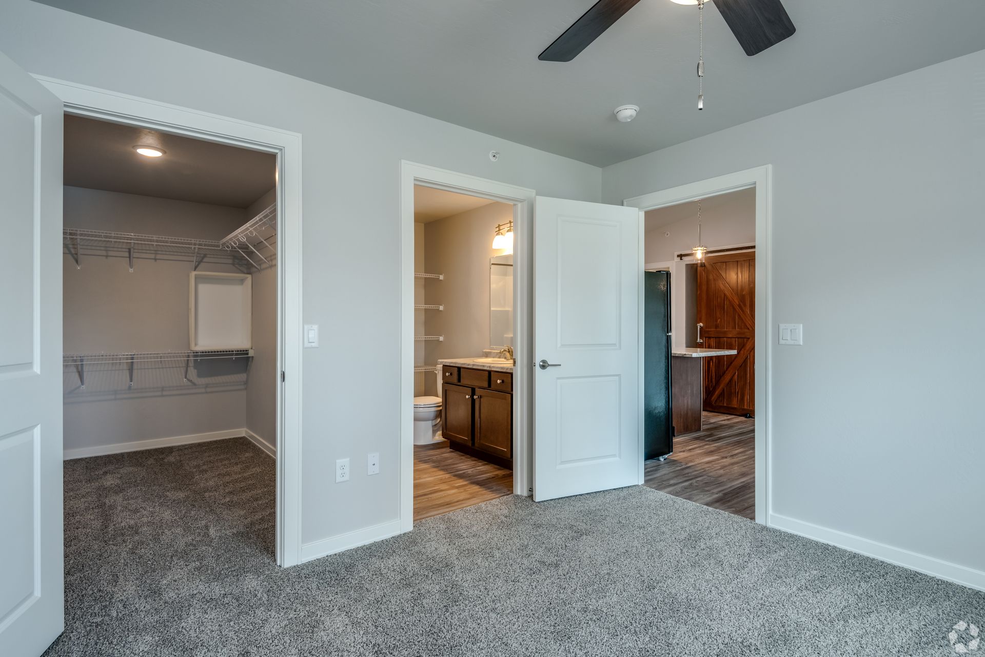 Spacious bedroom with open closets, gray carpet, and a ceiling fan, leading to a bathroom and hallway.