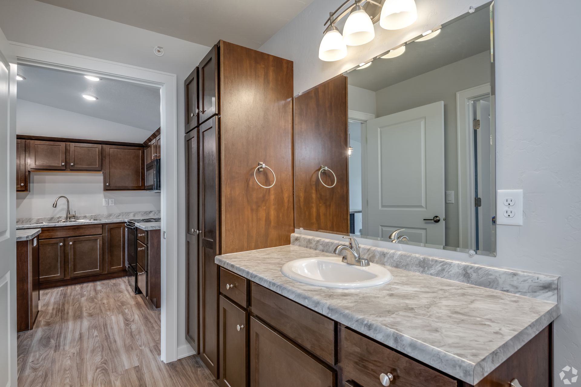 Bathroom vanity with marble countertop, mirror, and wood cabinets, opening to a kitchen area.