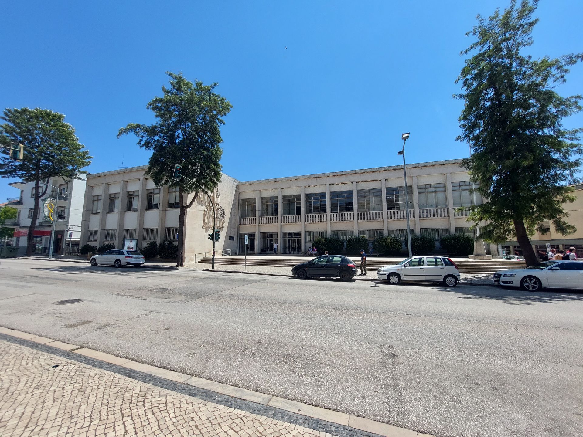 Um prédio de concreto de dois andares com várias janelas. Carros estacionados na rua em frente ao prédio, árvores em ambos os lados.